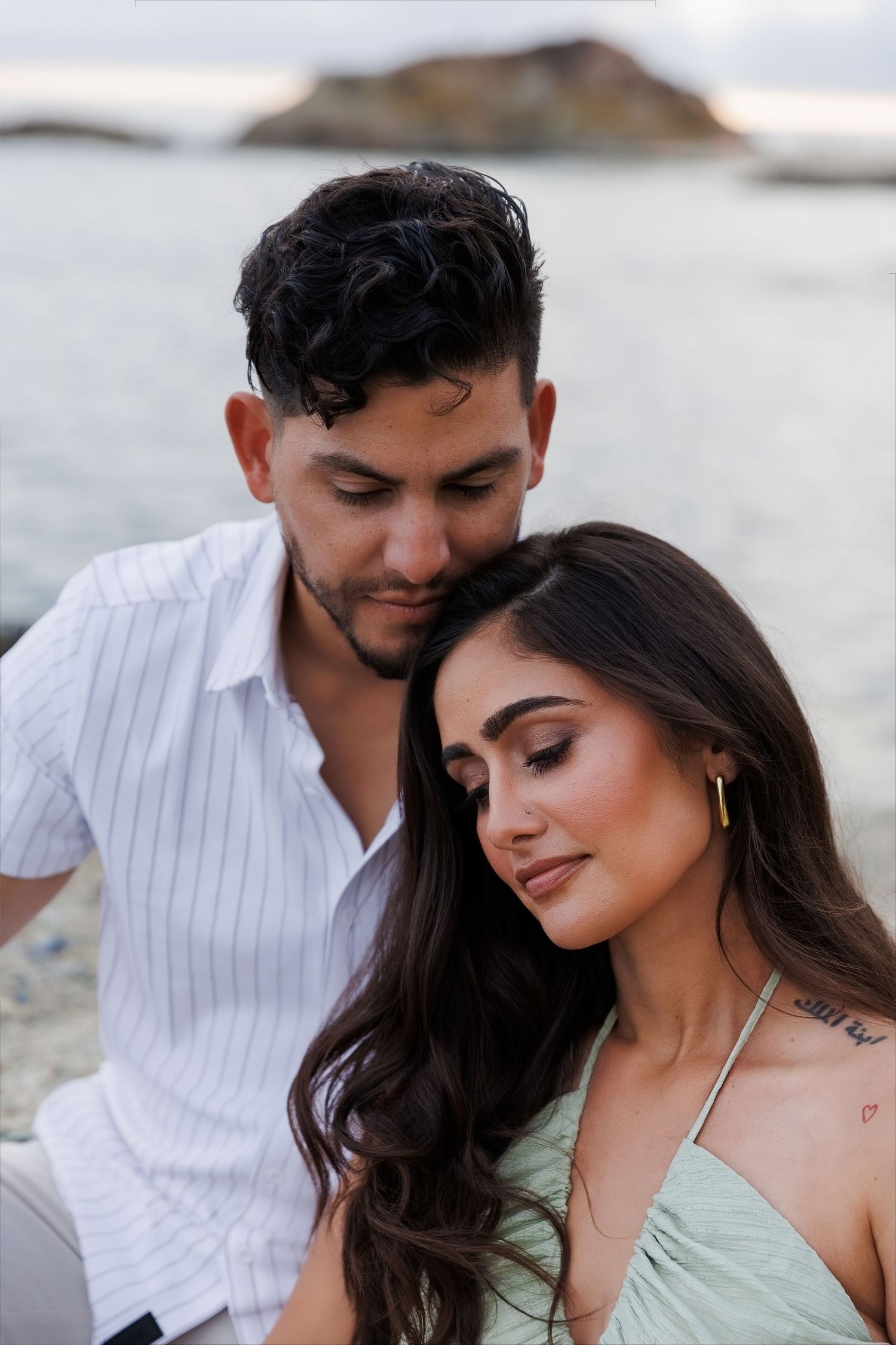 A romantic couple poses on a beach at dusk, woman in mint halter top rests head on mans shoulder.