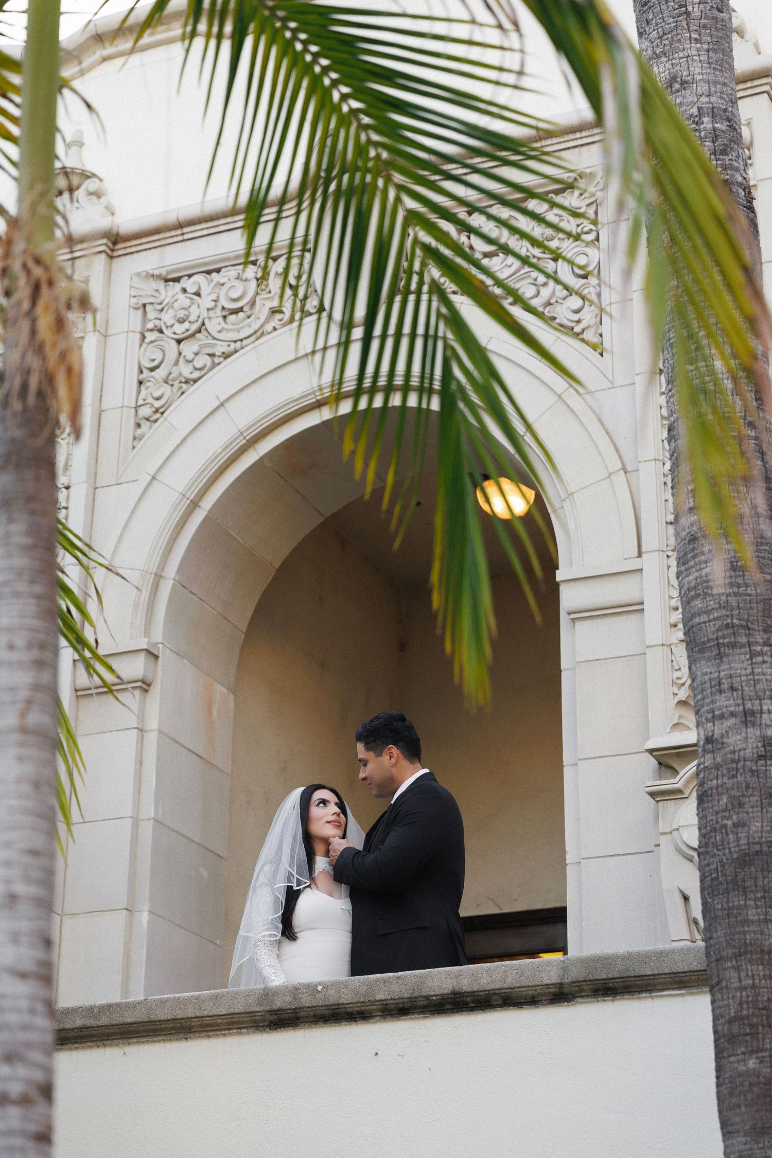 Bride and groom share a romantic moment under an ornate white arch, framed by tropical palm trees.