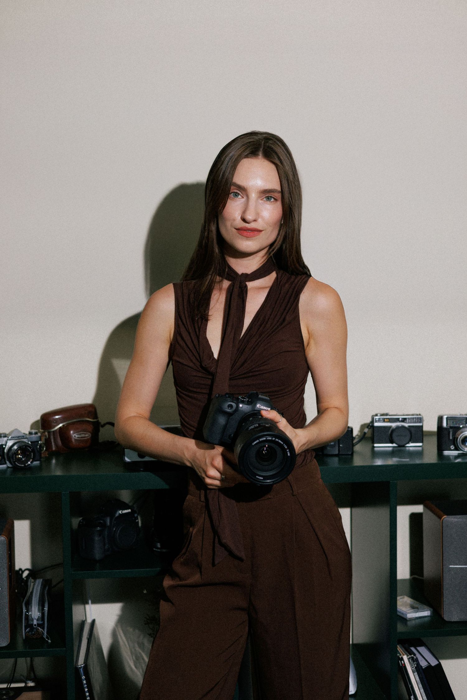 Woman in brown outfit holding a DSLR camera, posing in front of a shelf displaying vintage film cameras.