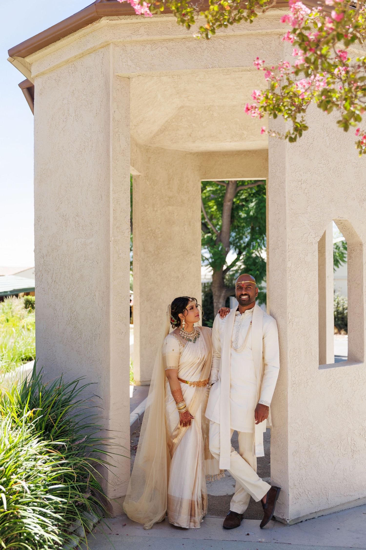 An Indian bride and groom in matching cream outfits pose together under an archway at their wedding venue.
