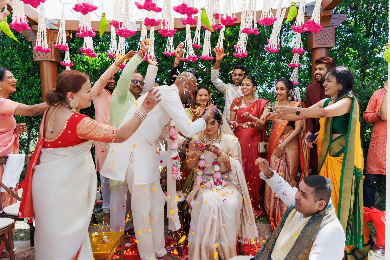 Colorful Indian wedding ceremony with guests tossing flower petals over the bride and groom outdoors.