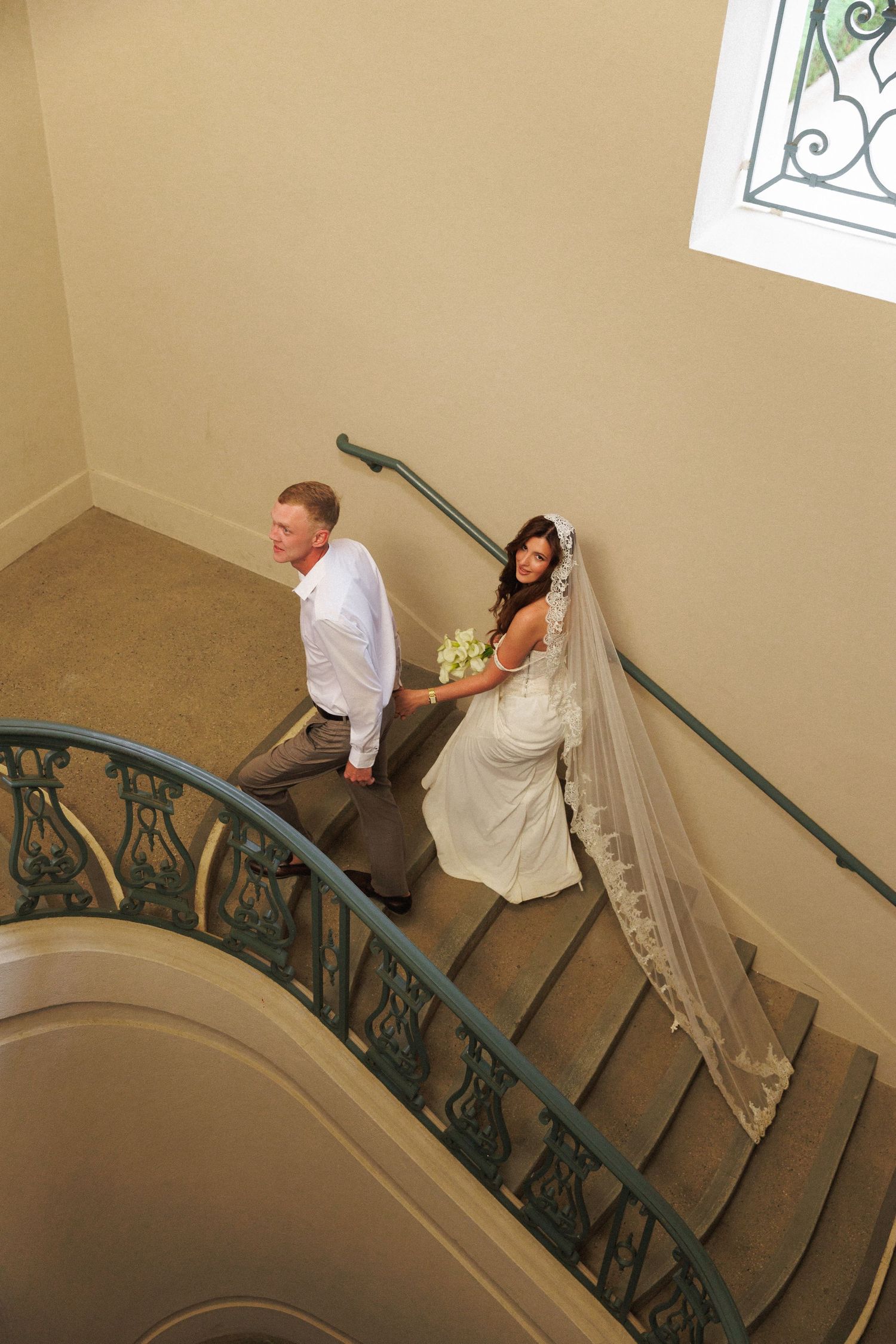 Bride and groom walking up an elegant curved staircase, bride holding bouquet with long veil trailing behind her.