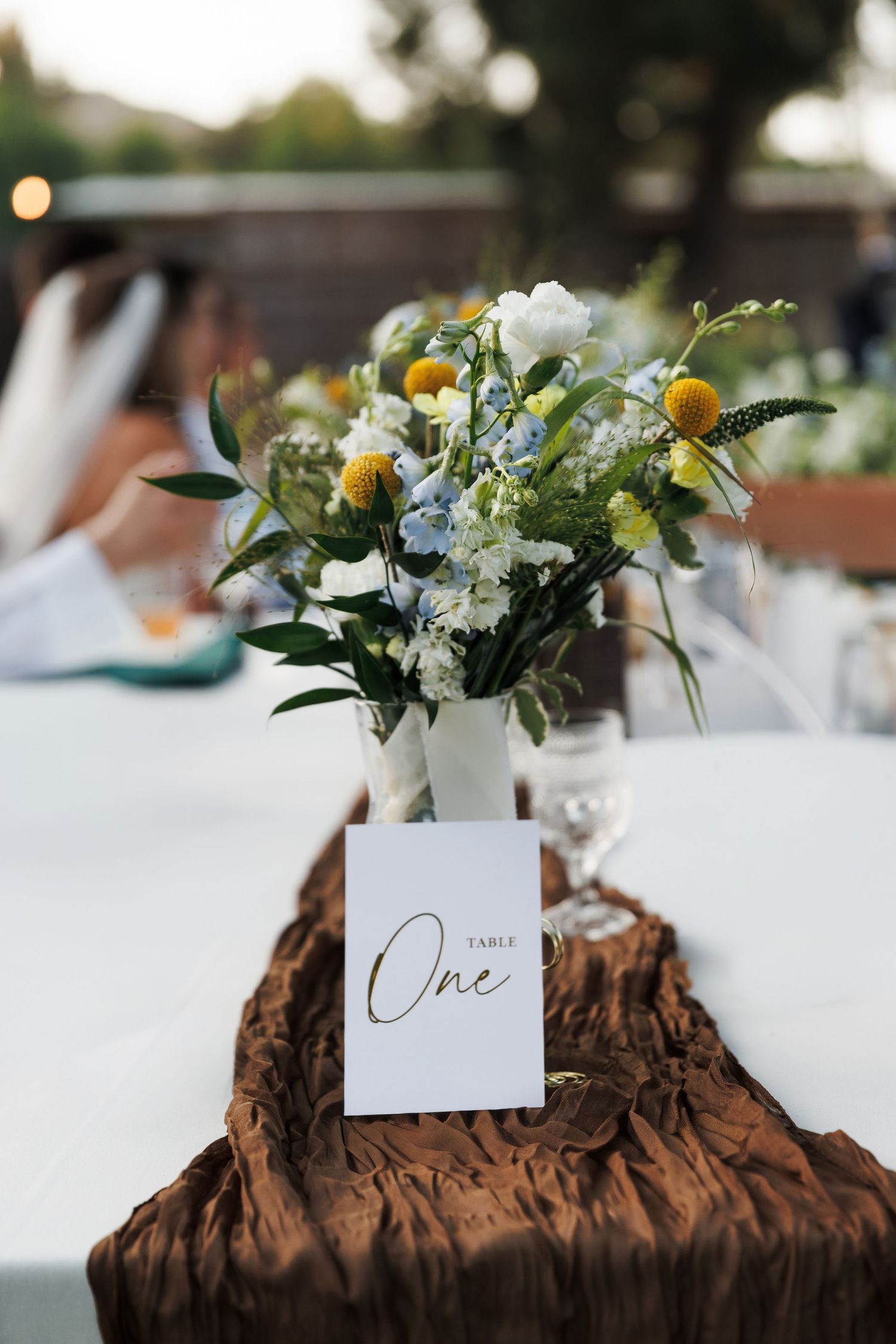 Wedding Table One sign with white vase of blue, yellow, and white flowers on brown linen tablerunner.