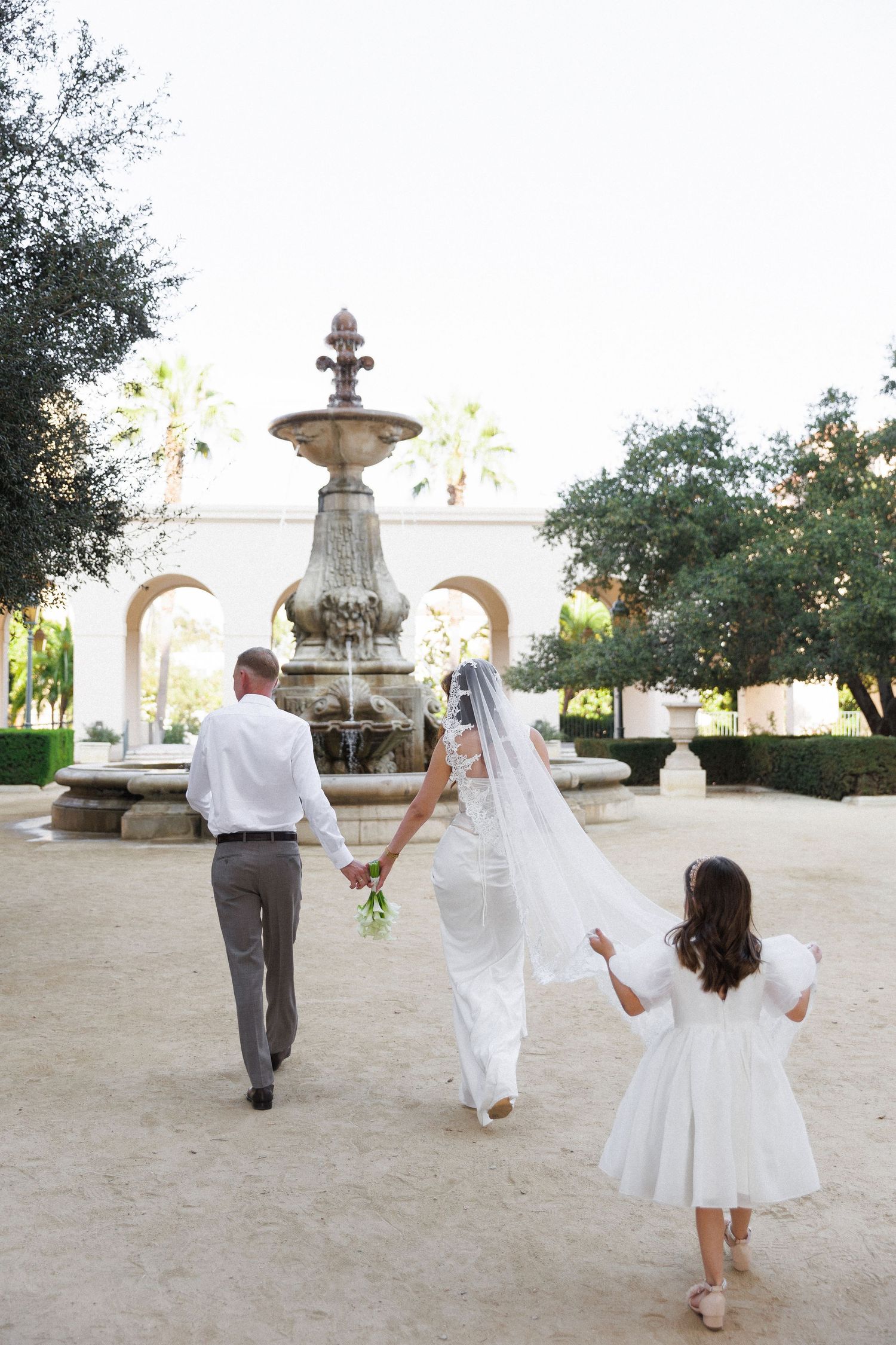 Bride and groom walking hand in hand with flower girl toward ornate stone fountain in elegant garden wedding venue.