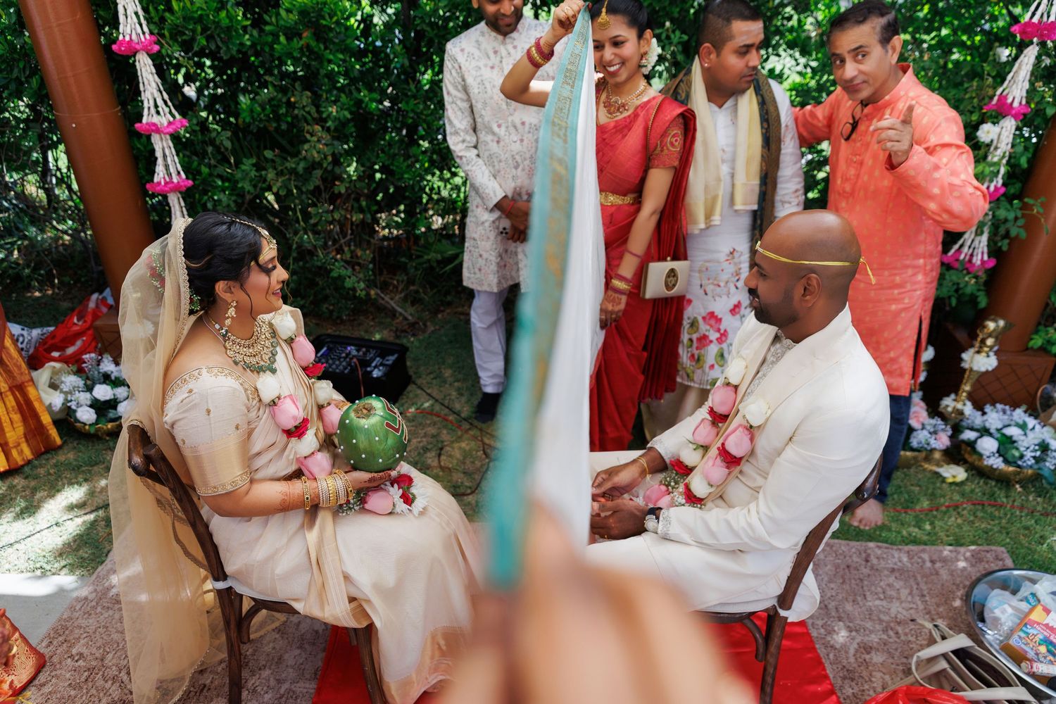 Indian wedding ceremony with bride and groom seated outdoors, surrounded by guests in traditional red and gold attire.