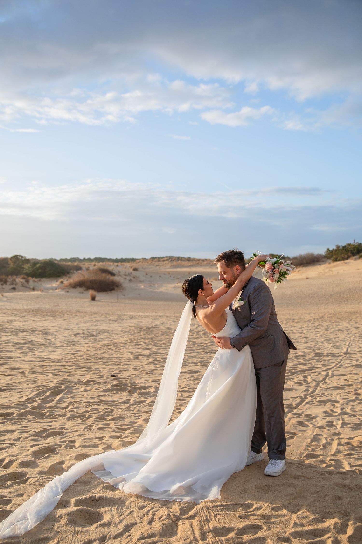 A couple shares a romantic moment on a sandy beach with flowing white dress and gray suit against cloudy sky at sunset.