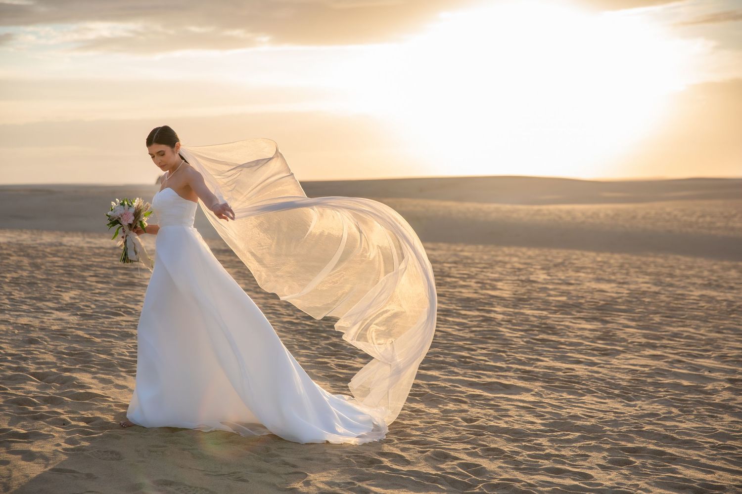 A couple in wedding attire stands on a sandy beach at sunset as a white veil flows gracefully in the wind.