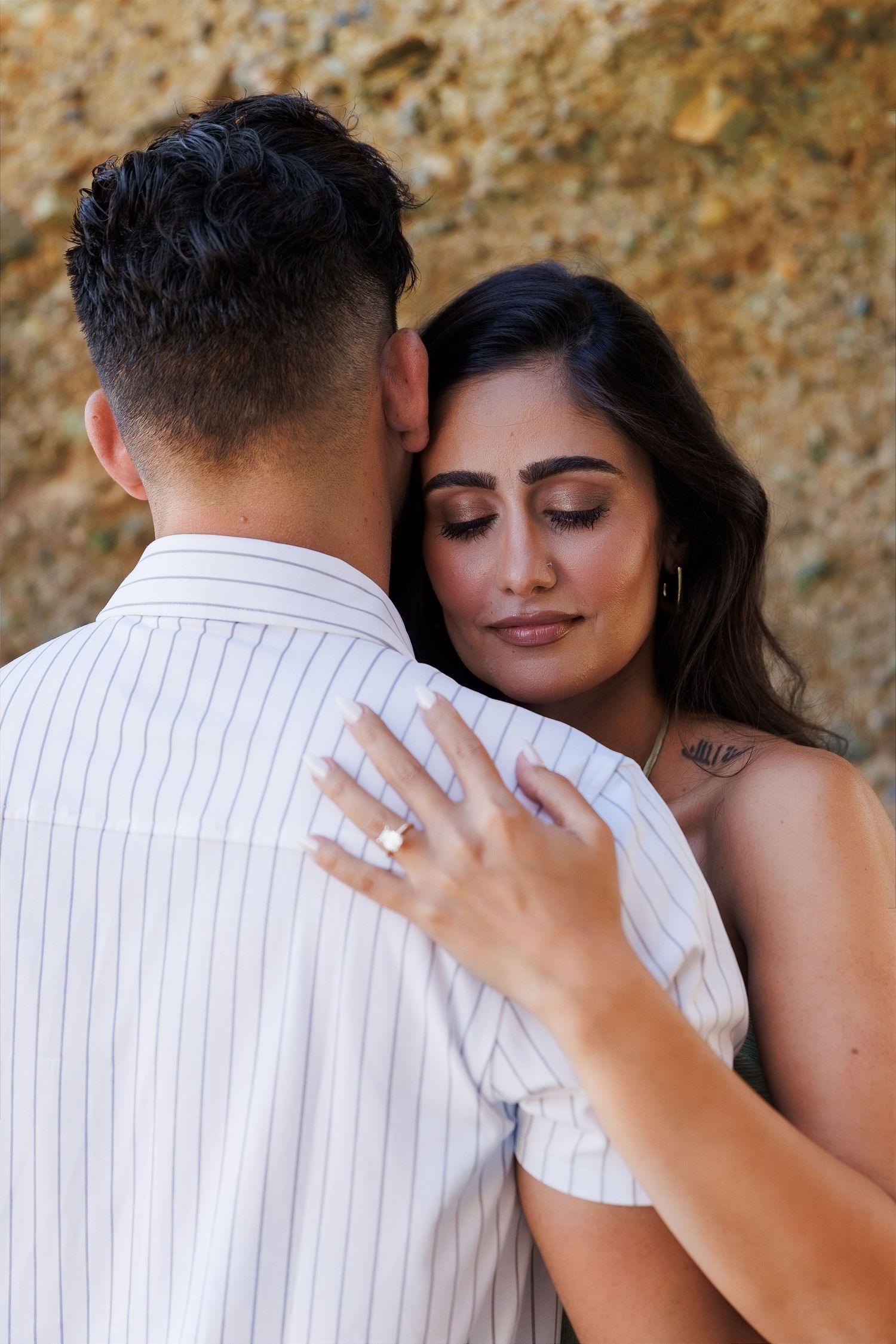 Couple embracing tenderly, woman with engagement ring and tattoo resting head on mans shoulder outdoors.