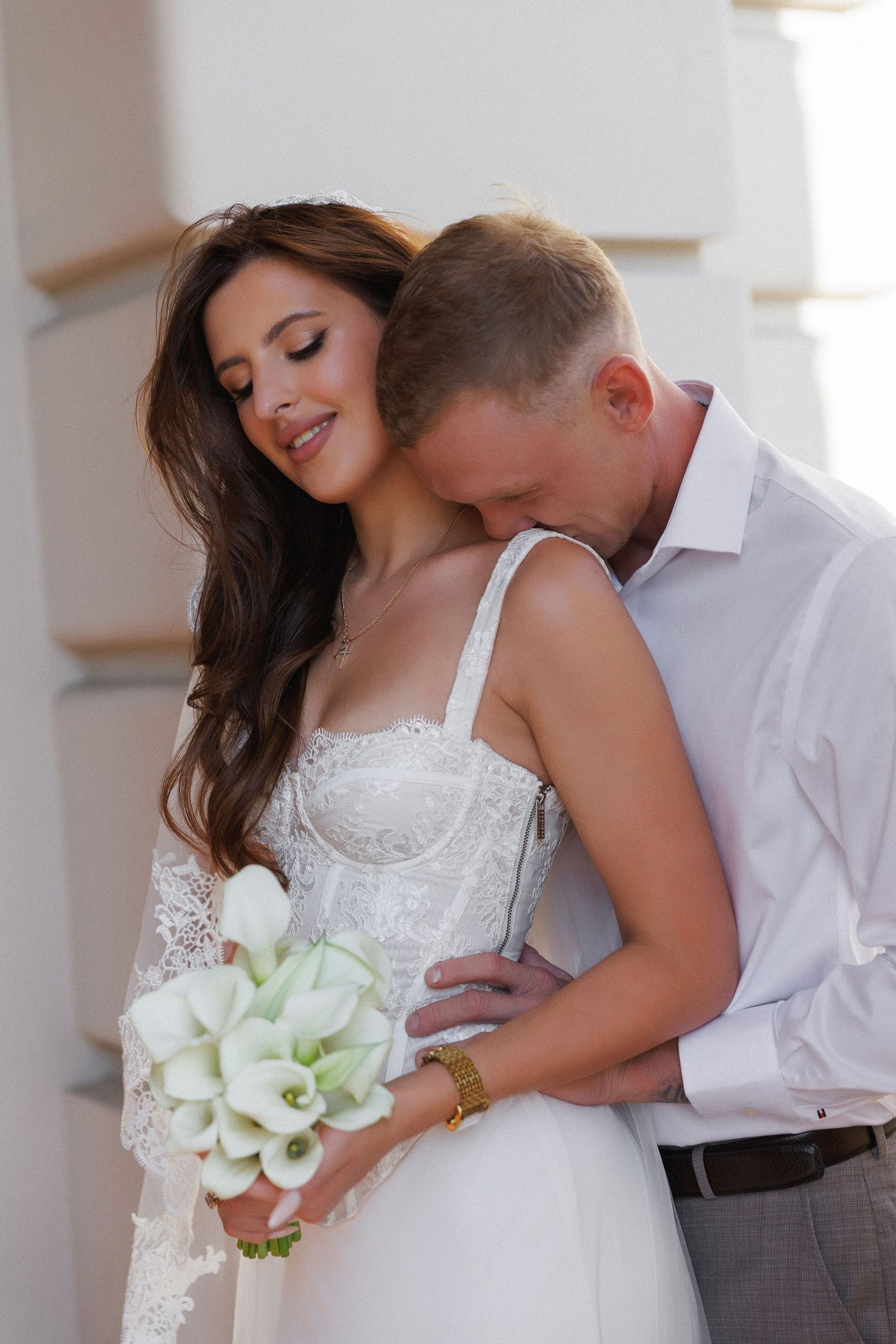 Bride in white lace dress holding calla lily bouquet, groom in white shirt embracing her from behind outdoors.