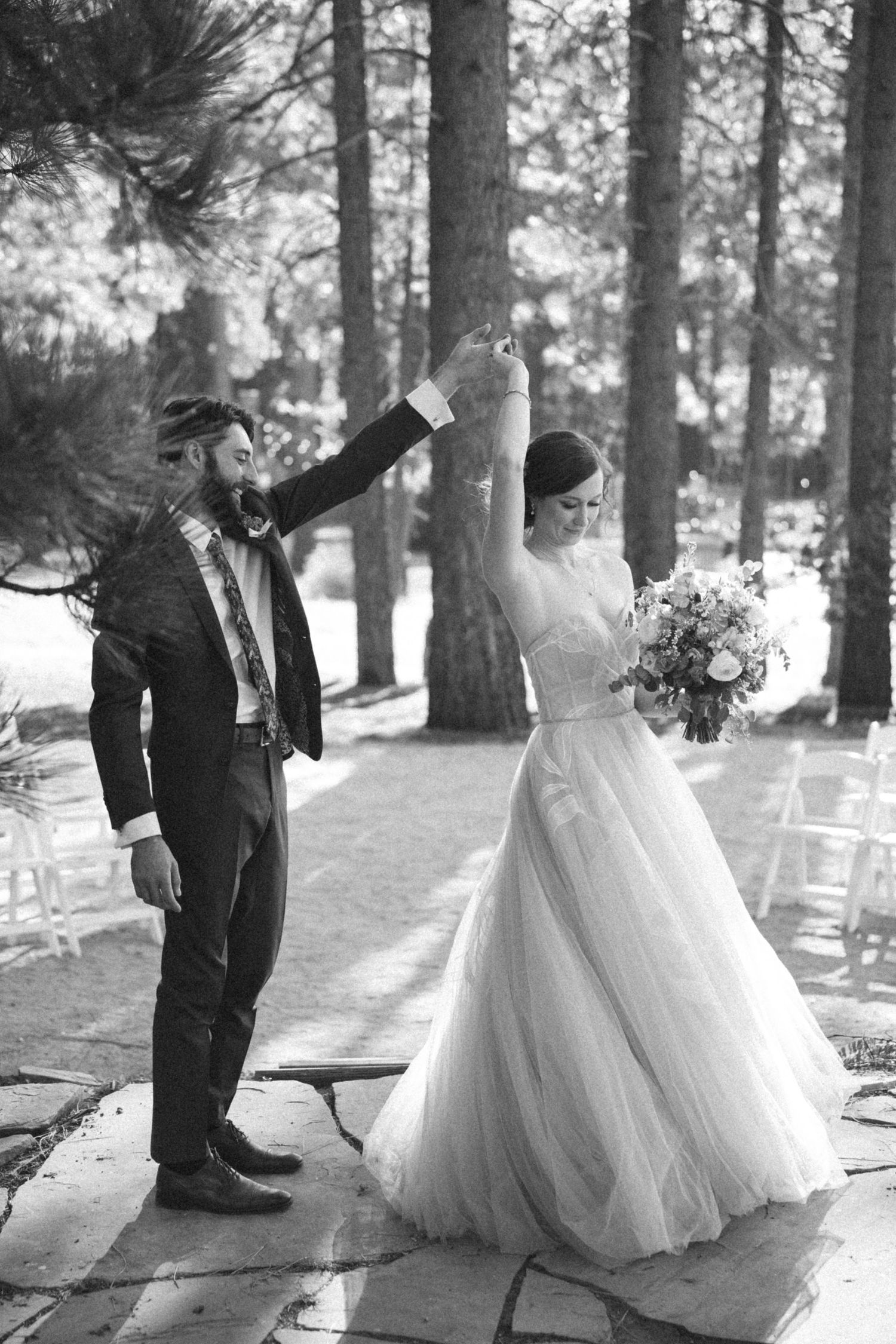 Black and white photo of a groom spinning his bride in a wedding dress outdoors among tall pine trees.