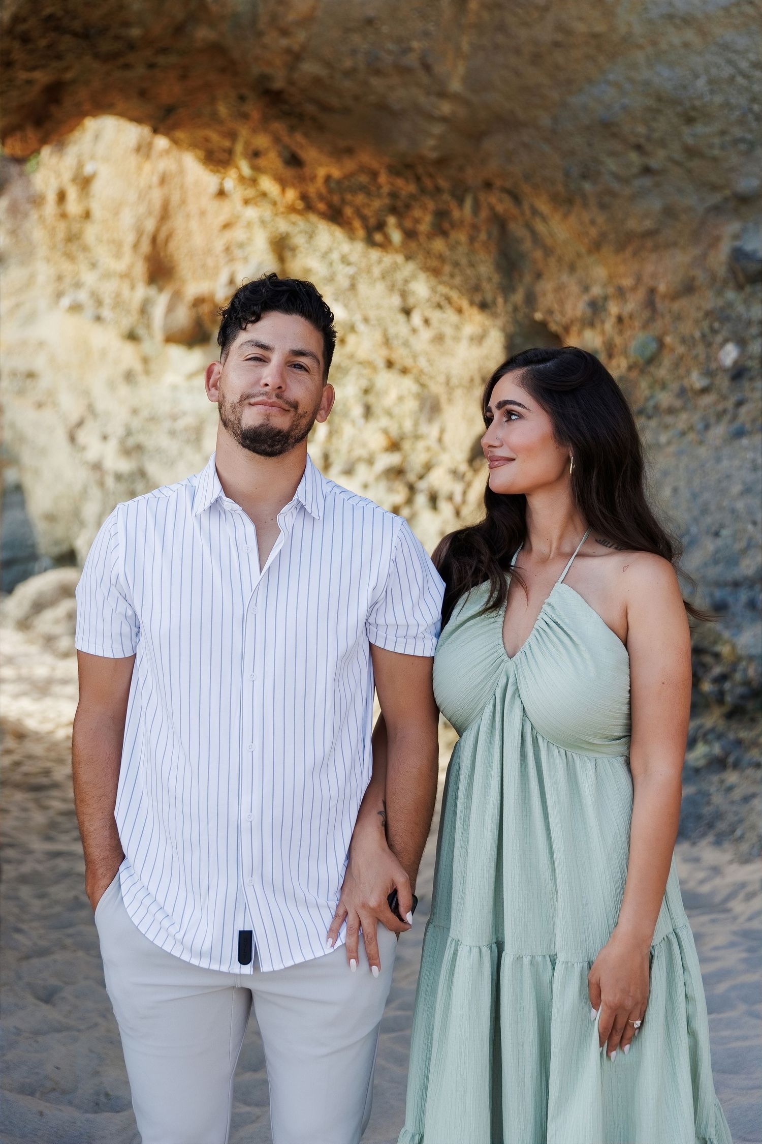 A couple holds hands on a beach, man in striped shirt and woman in sage green dress, with rocky cliffs behind.