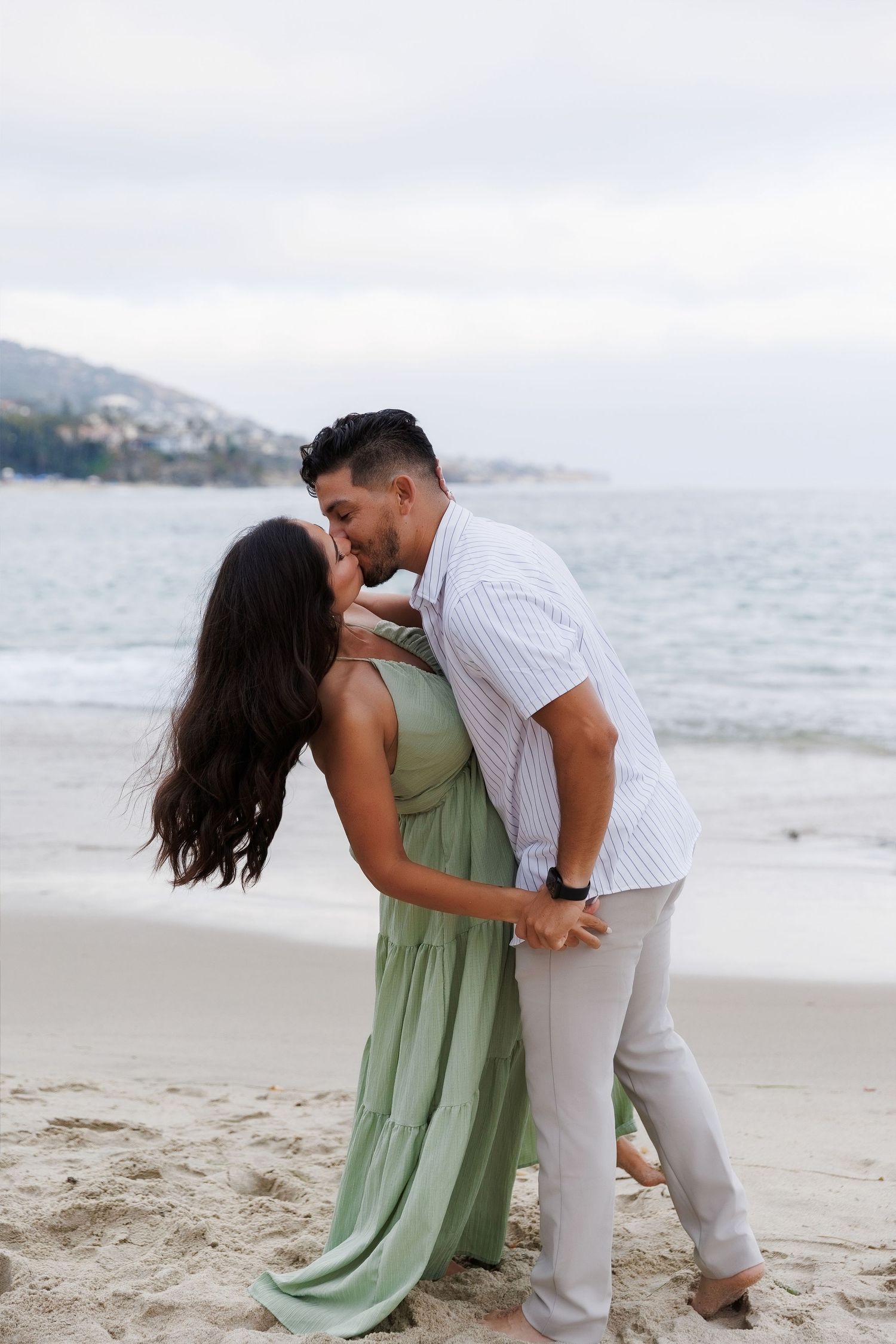Couple sharing a romantic kiss on a sandy beach, woman in green maxi dress, man in white striped shirt.