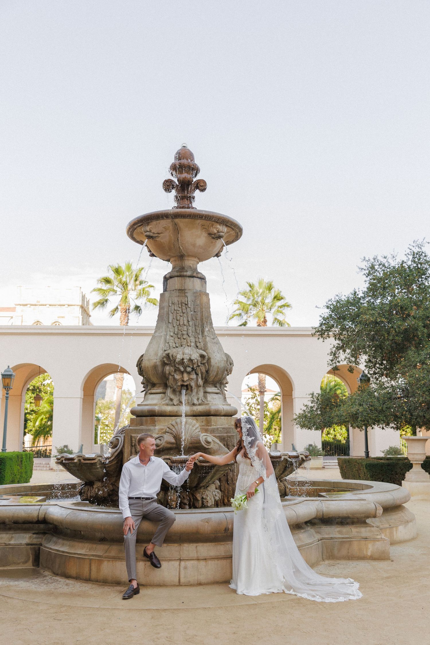 Bride and groom posing by an ornate stone fountain in a Mediterranean garden with palm trees and arched colonnade.