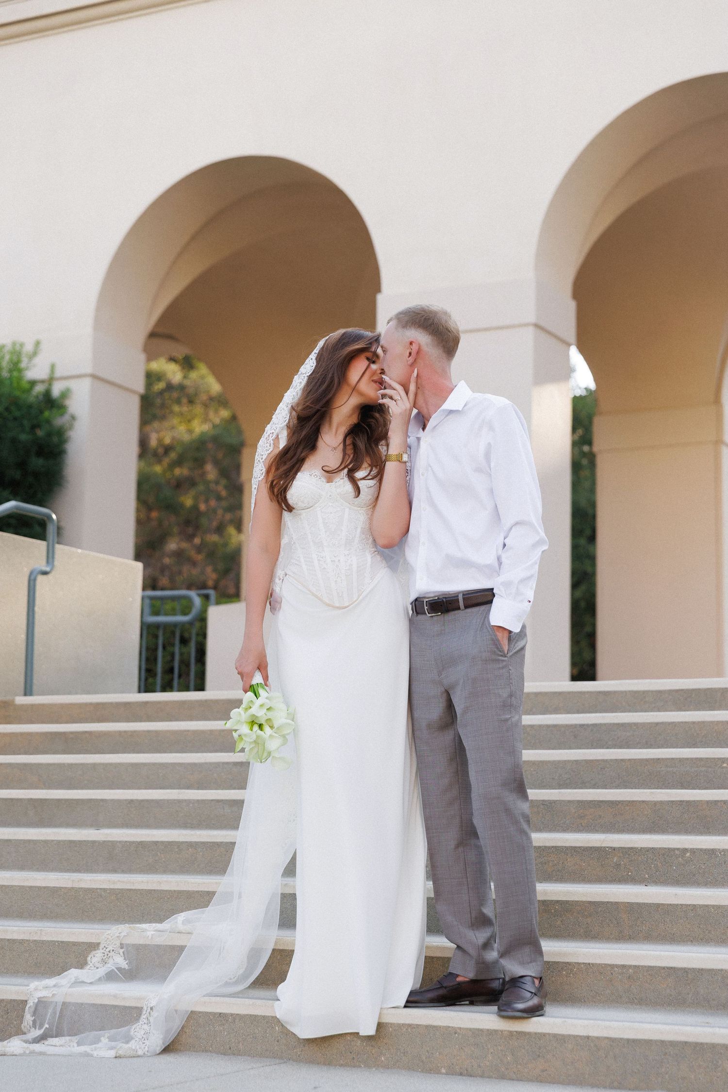 Bride and groom share a kiss on elegant stone steps with arched columns, bride holding white bouquet in wedding dress with veil.