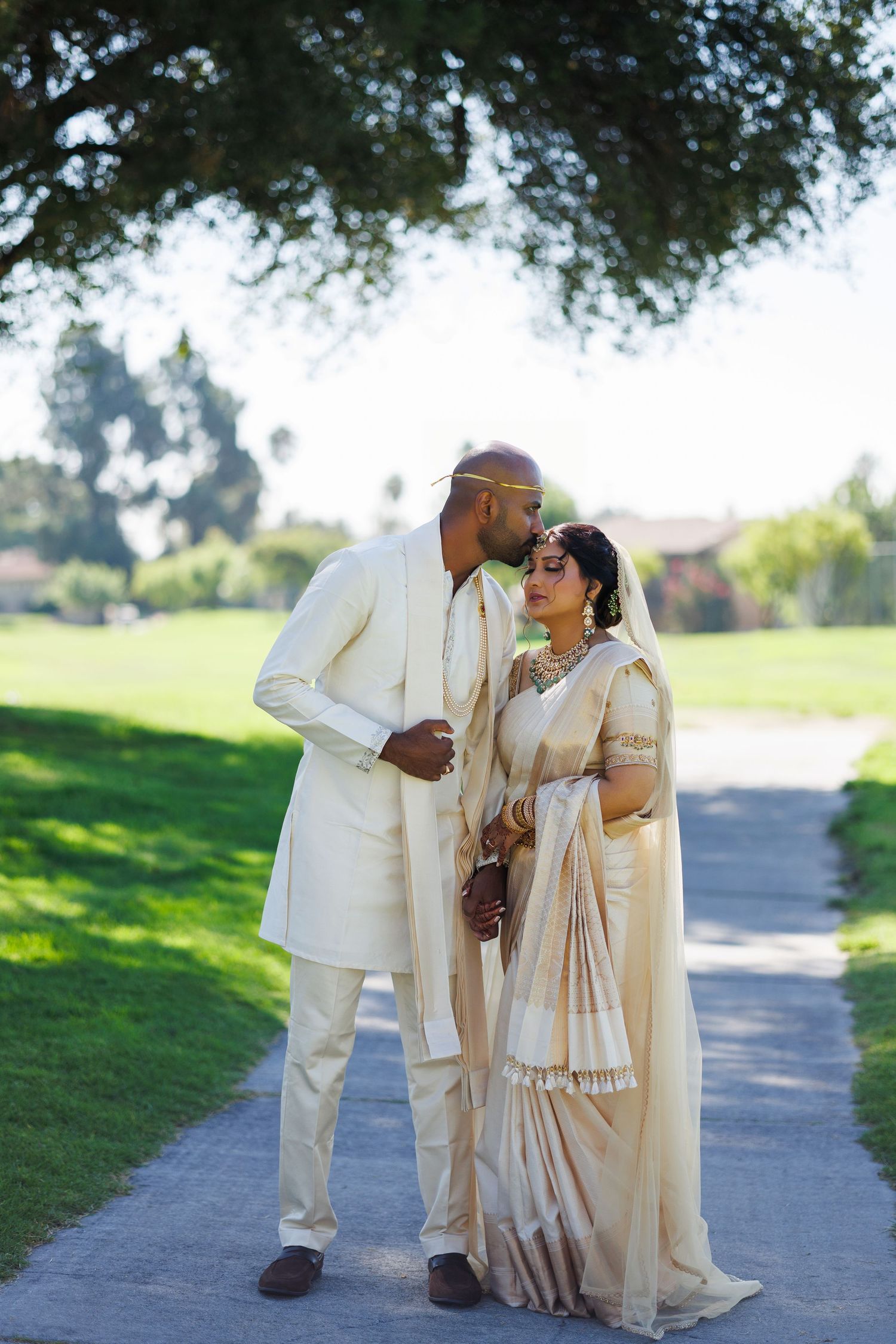 Indian couple in cream wedding attire sharing a romantic moment on a garden path, groom kissing bride's forehead.