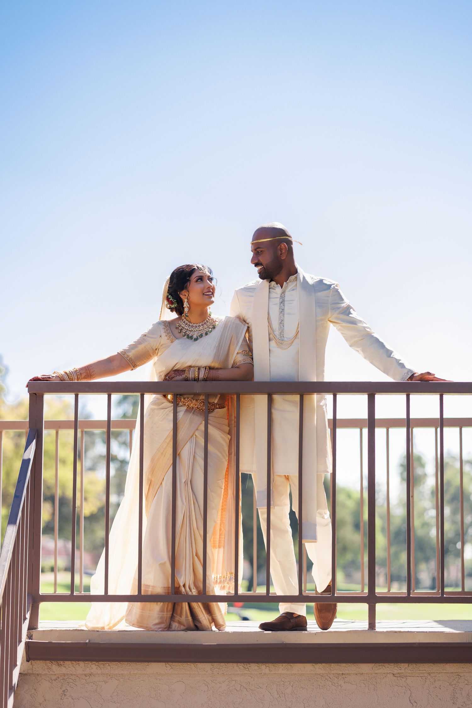 Indian couple in traditional white wedding attire standing on a balcony, sharing a romantic glance outdoors.