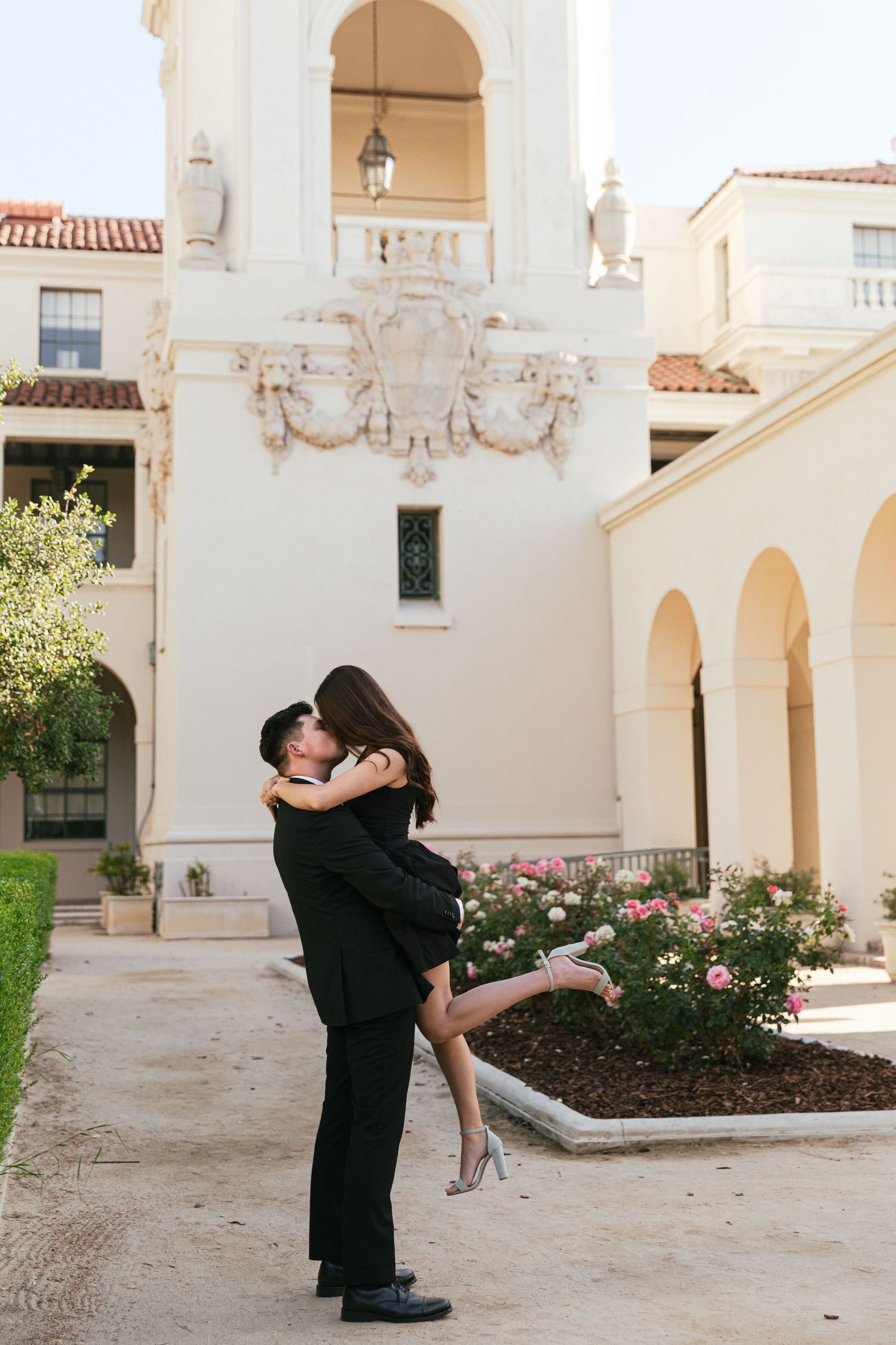 A romantic engagement photoshoot in front of a Spanish colonial style building with white arches and terra cotta tiles.