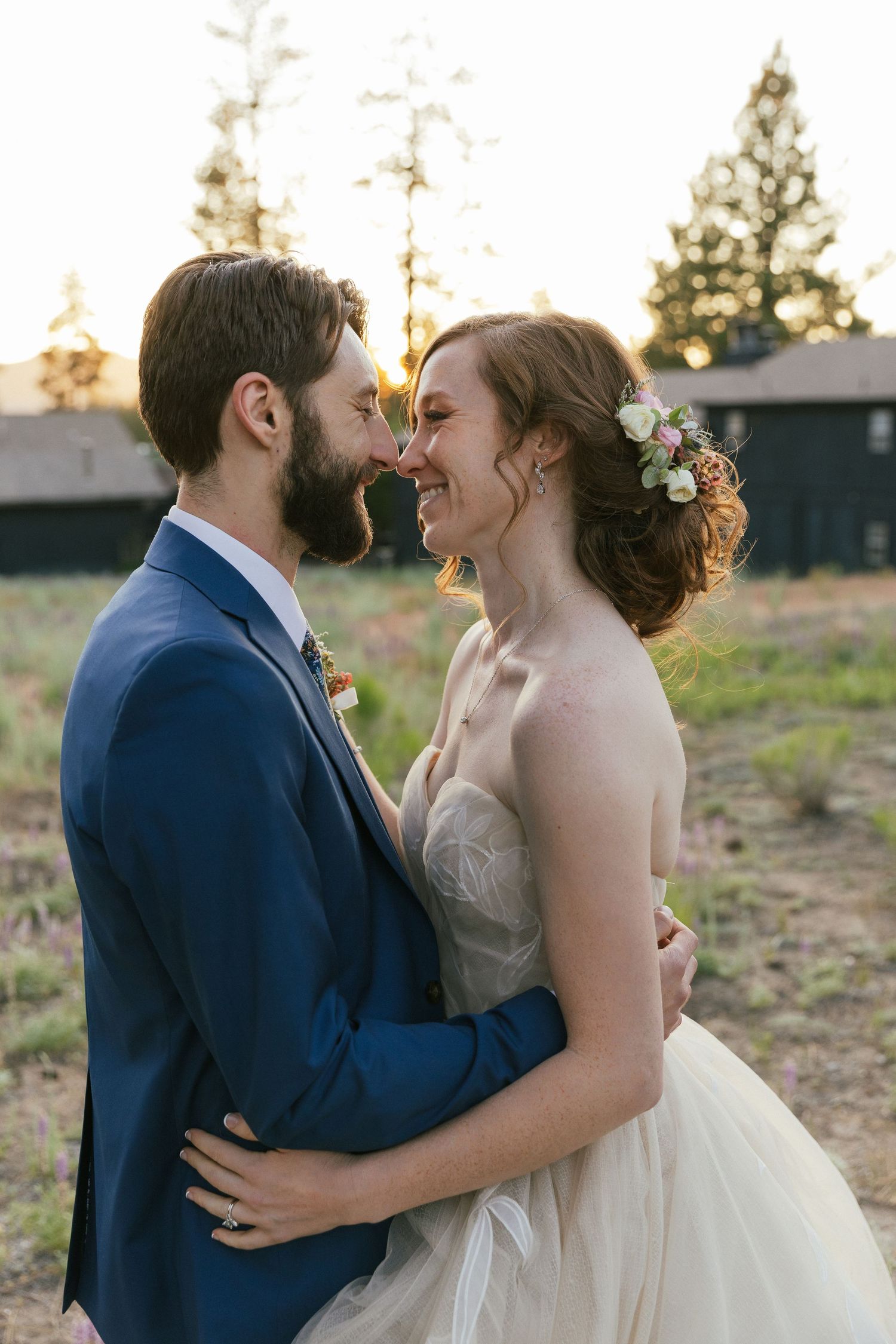 Bride and groom share a romantic sunset moment, smiling close together outdoors at a rustic wedding venue.