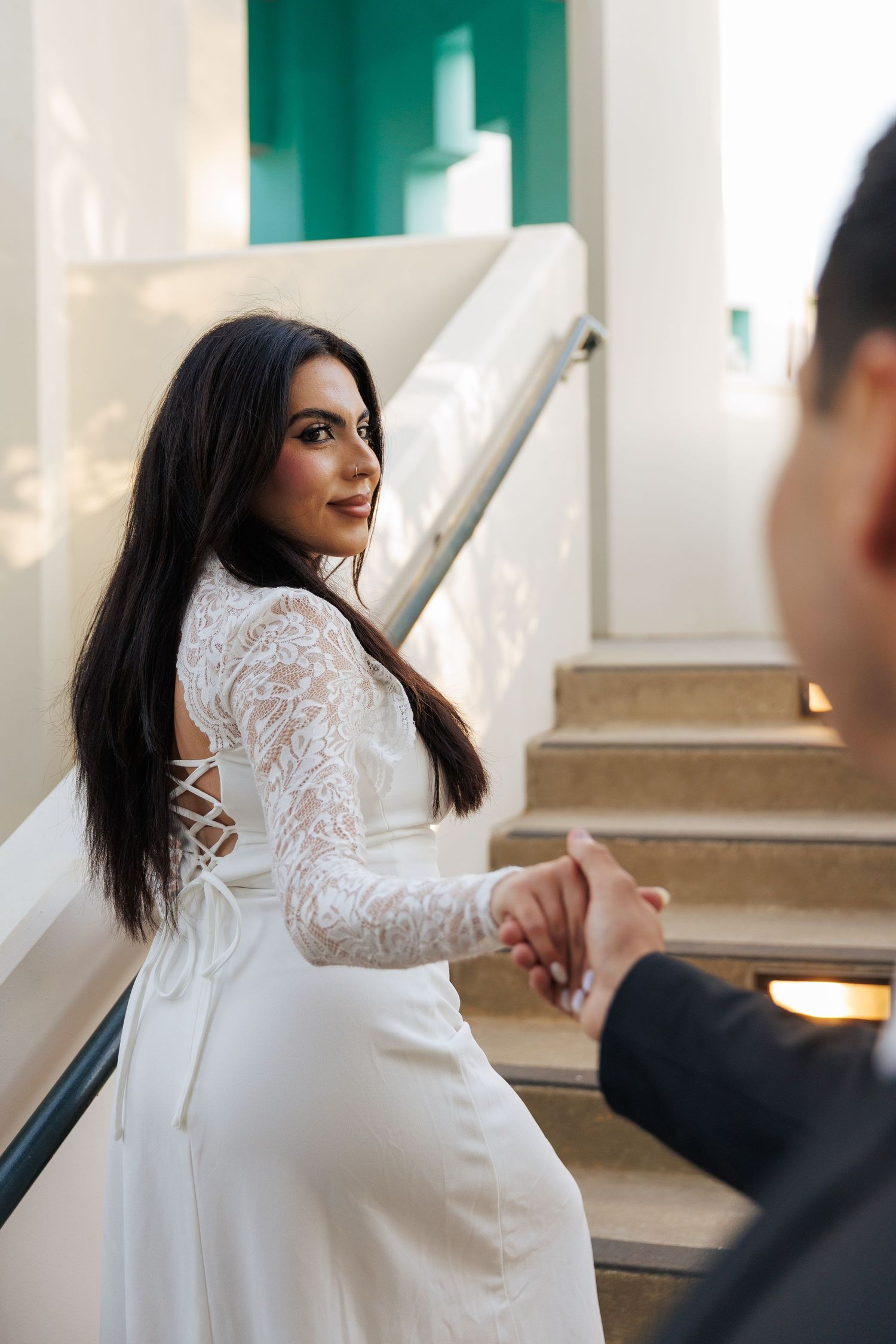Bride in white lace gown holds hands with groom while looking back on elegant staircase.