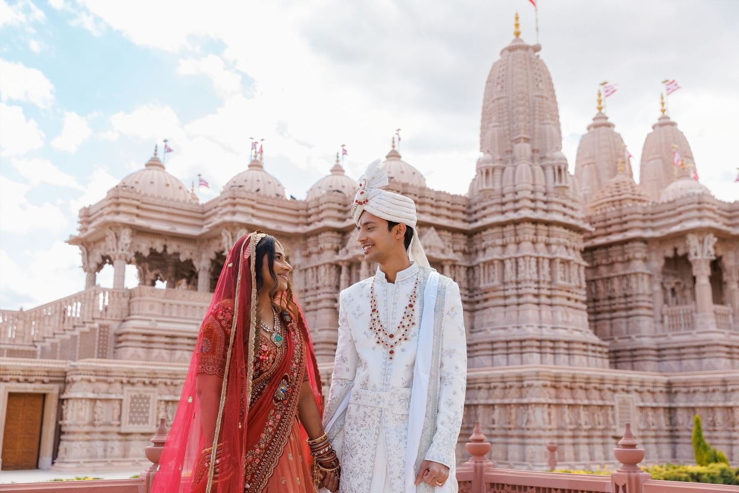Indian couple in traditional wedding attire holding hands before ornate BAPS Swaminarayan temple.