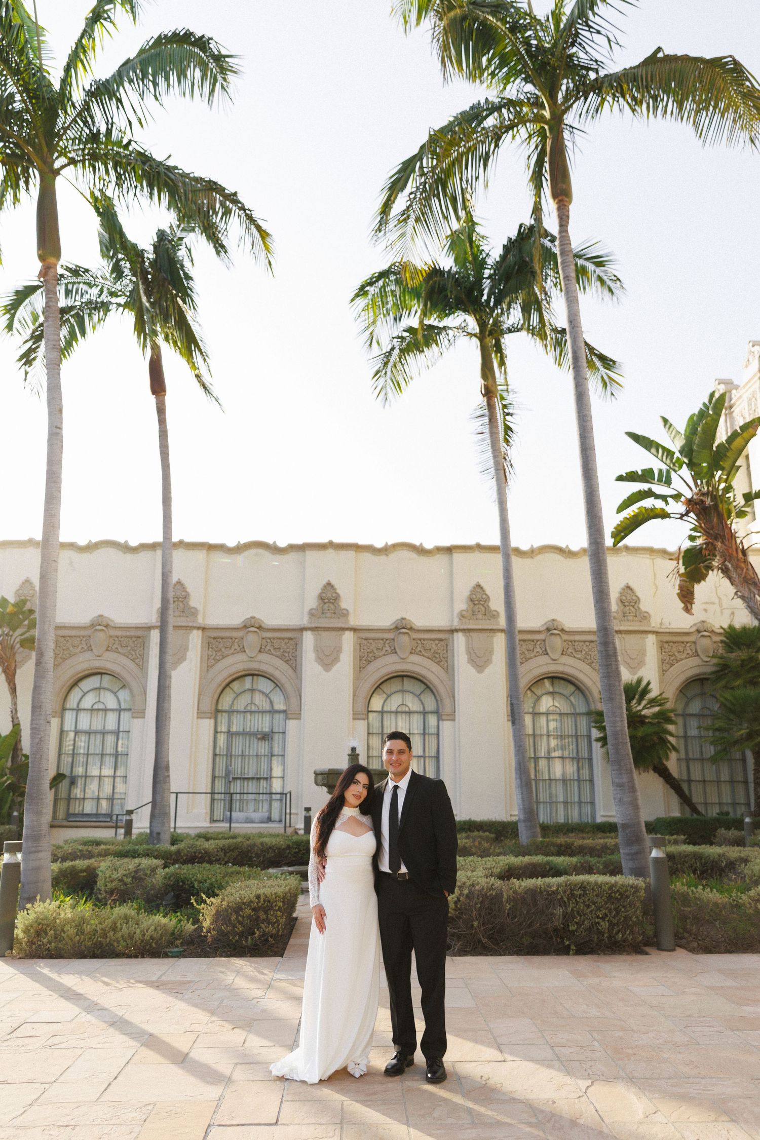 Bride and groom pose before a historic building surrounded by tall palm trees on their wedding day.