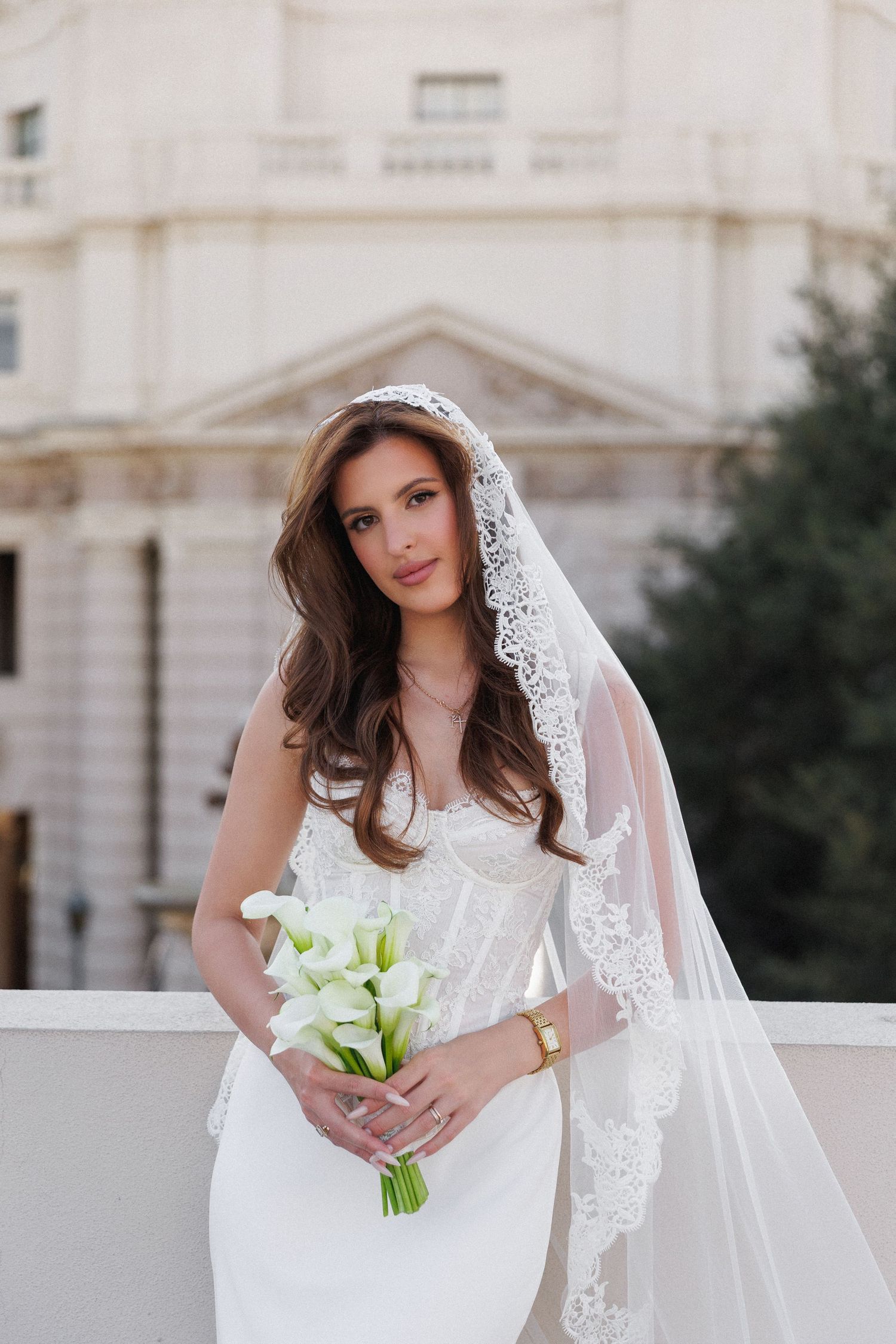 Elegant bride in white corset gown and lace veil holding calla lily bouquet, posing before a classical building.