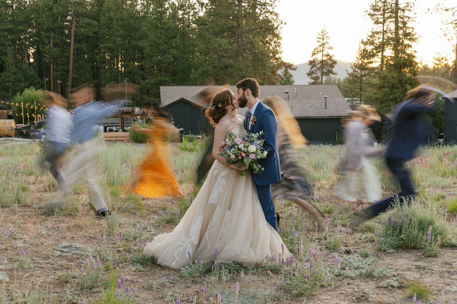 Bride and groom share a kiss outdoors at golden hour while blurred wedding guests move around them.