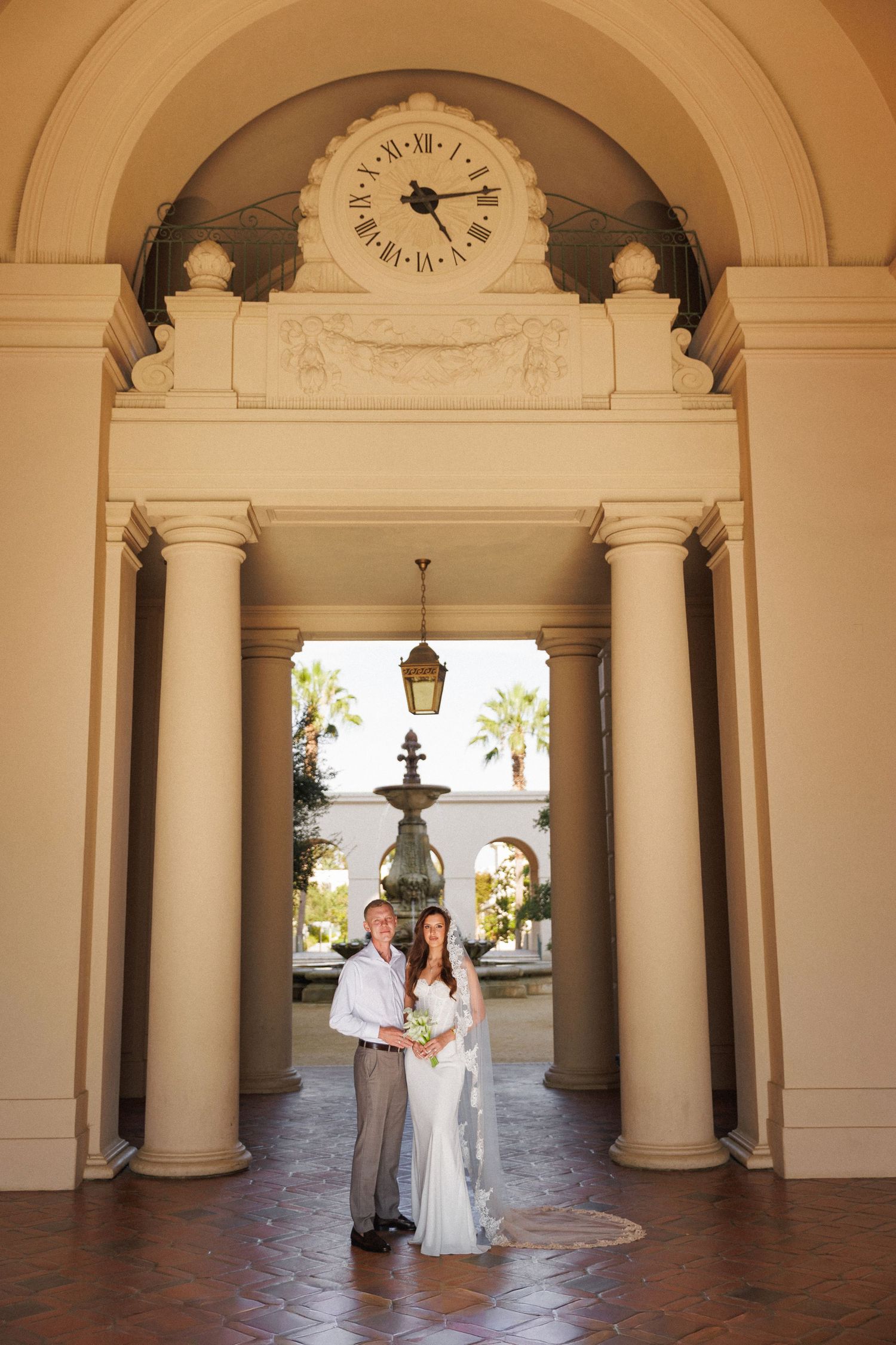 Bride and groom posing under elegant arched colonnade with clock and fountain at luxury wedding venue.