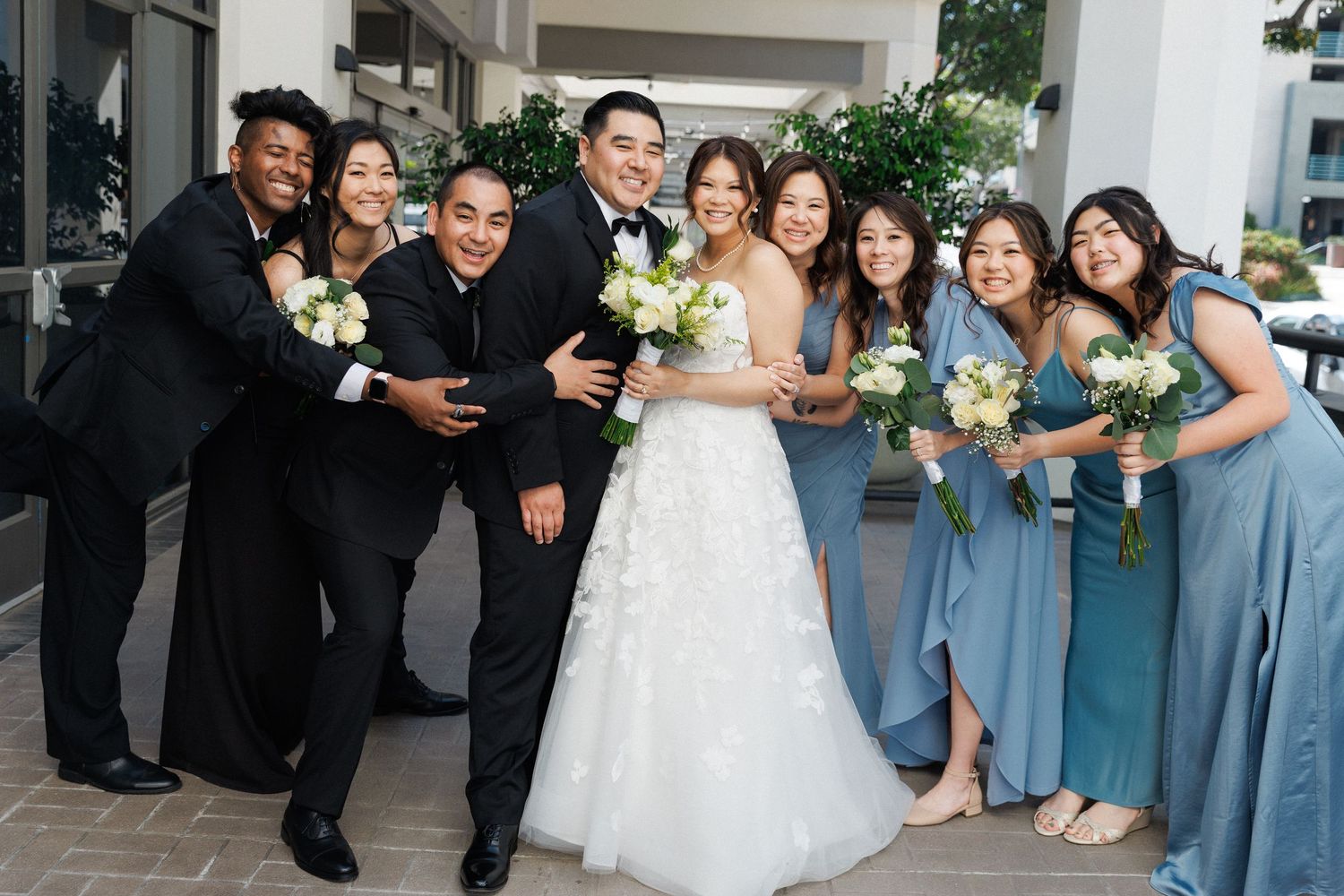 Wedding party poses together outdoors, bride in white gown with groom in tuxedo surrounded by bridesmaids in blue dresses.