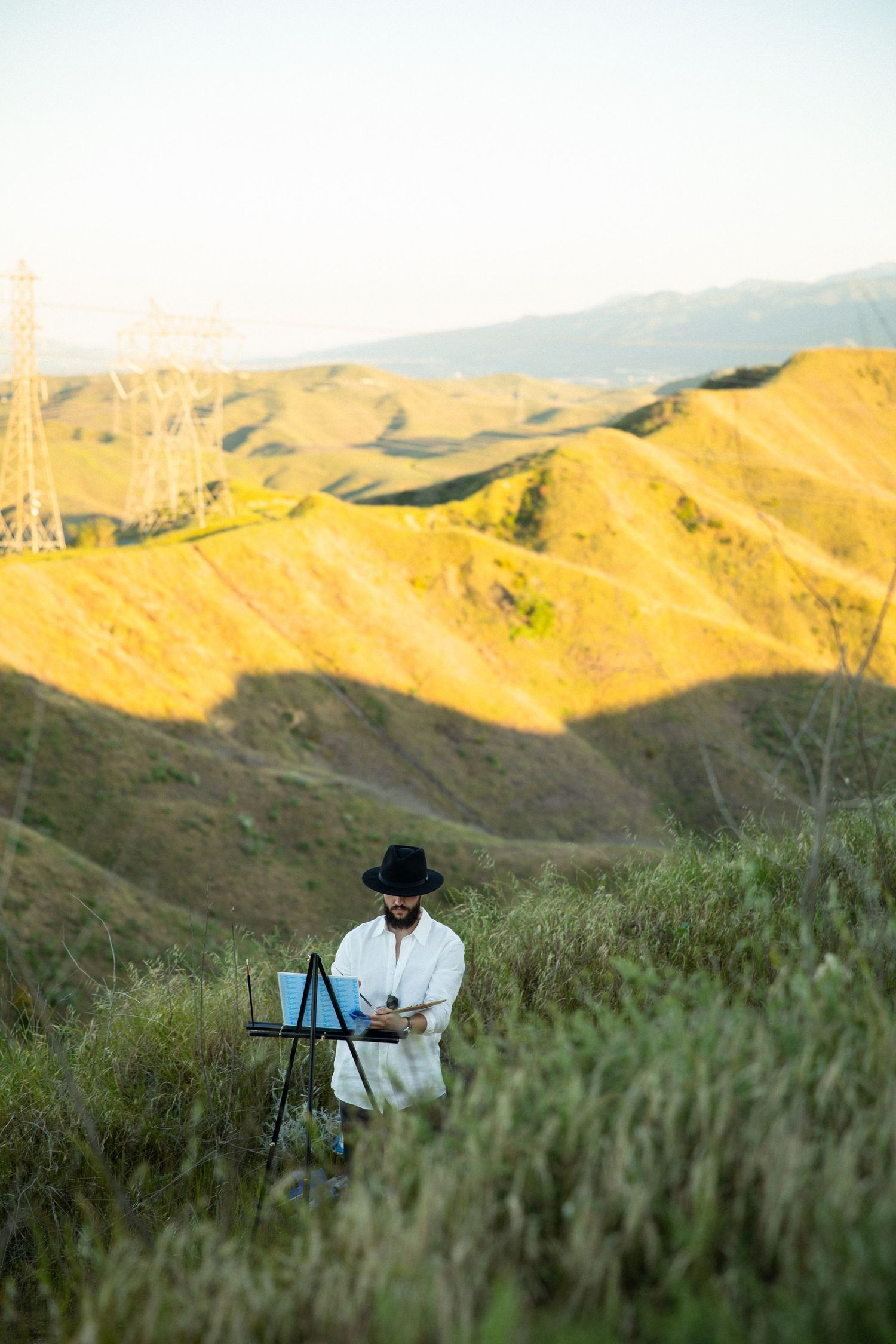 Artist in a black hat painting on an easel against golden rolling hills at sunset.