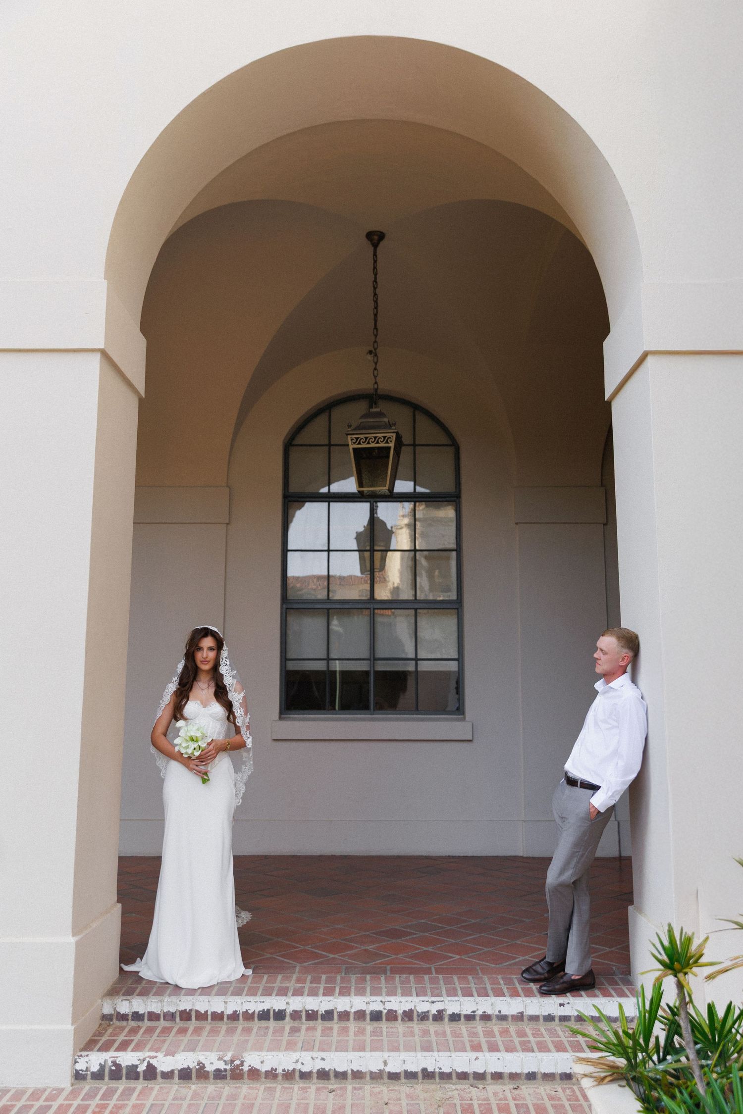 Bride in white gown and groom in white shirt stand apart under elegant arch with hanging lantern.