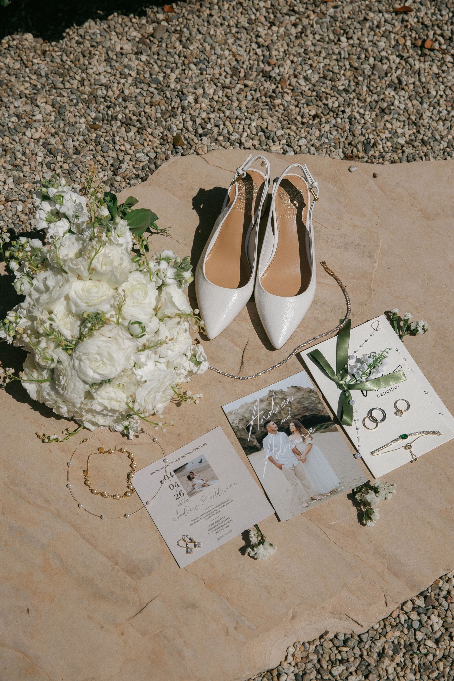 Bridal flat shoes, white rose bouquet, wedding rings, and invitation suite styled on a stone surface.
