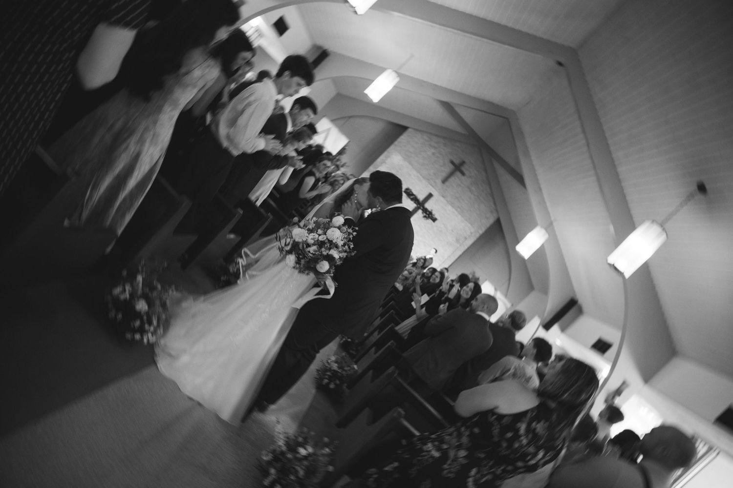 Black and white photo of a wedding ceremony inside a church with a cross on the wall and bridal party at the altar.