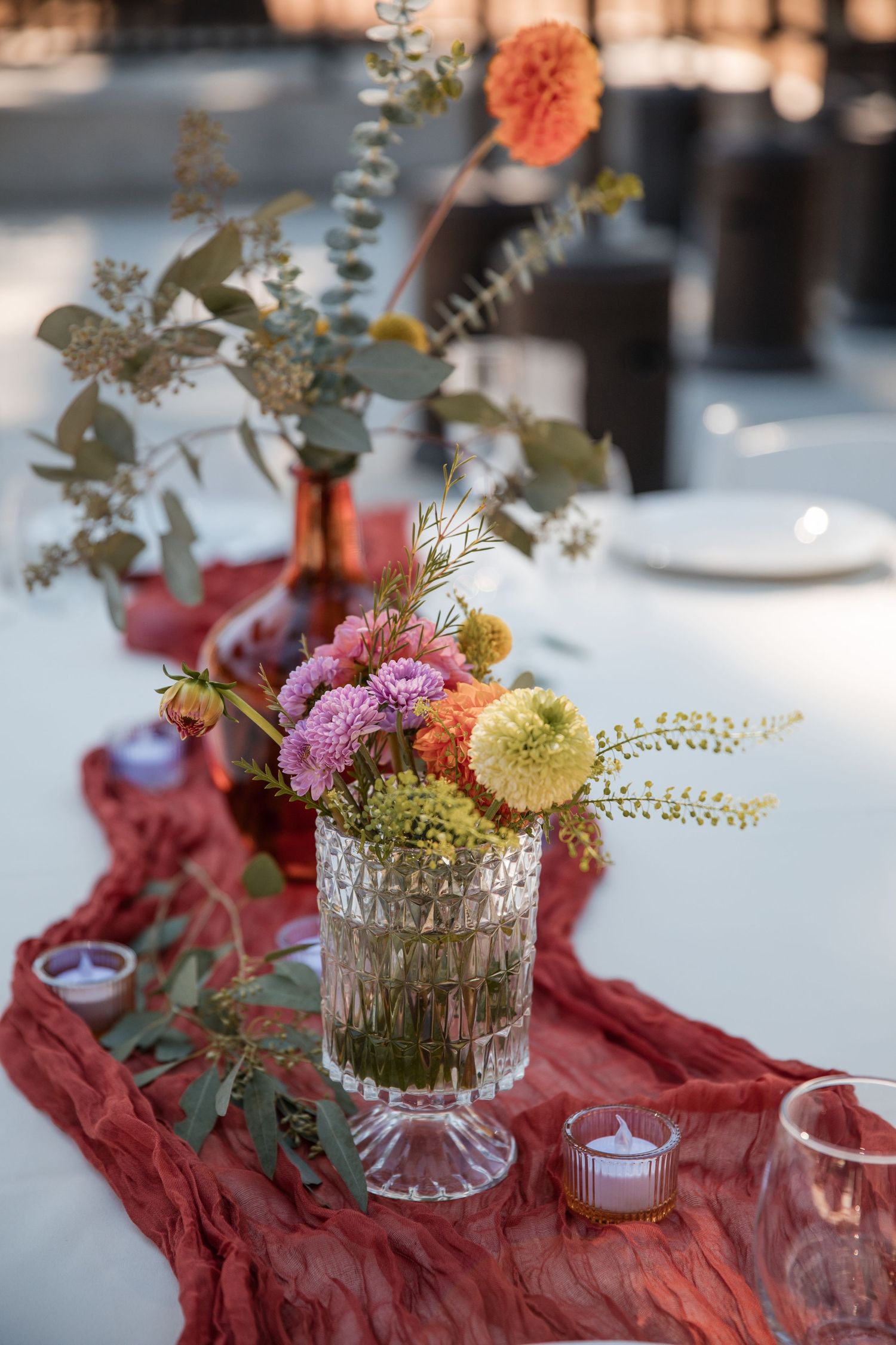 Delicate floral centerpieces with pink and yellow blooms sit in crystal vases on a flowing red table runner.