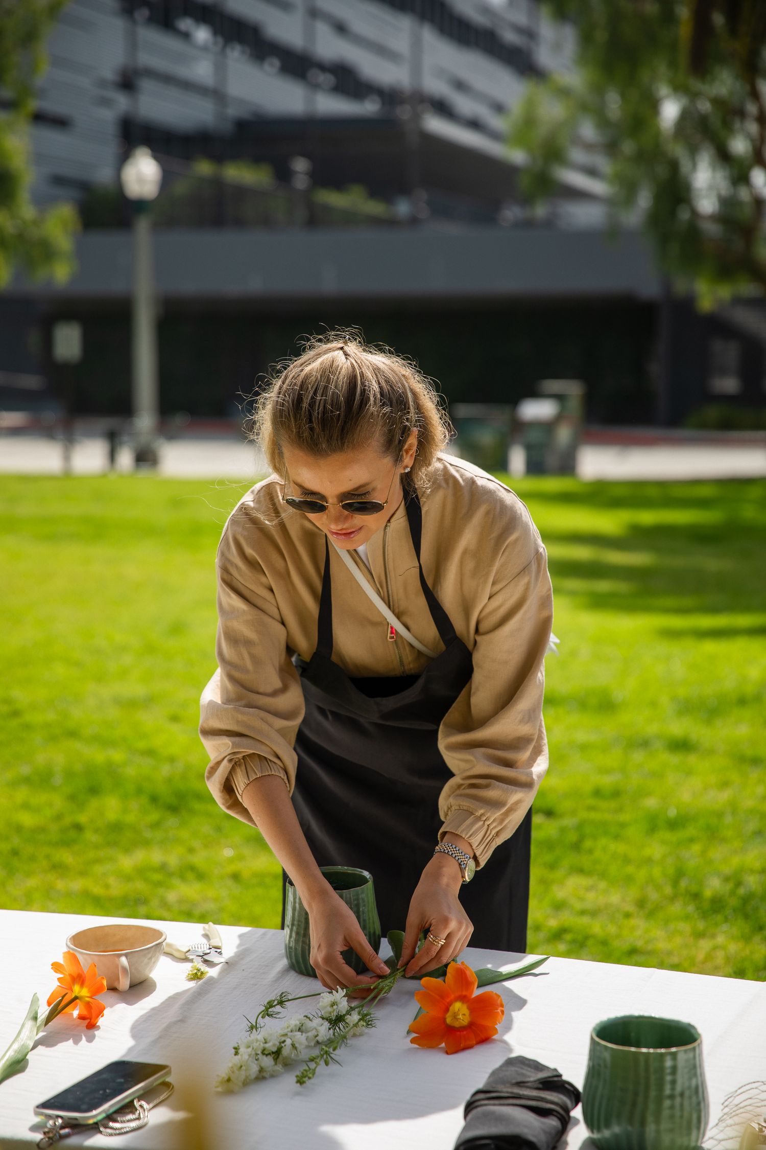 Person in beige sweater and black apron working on floral arrangements at an outdoor table.