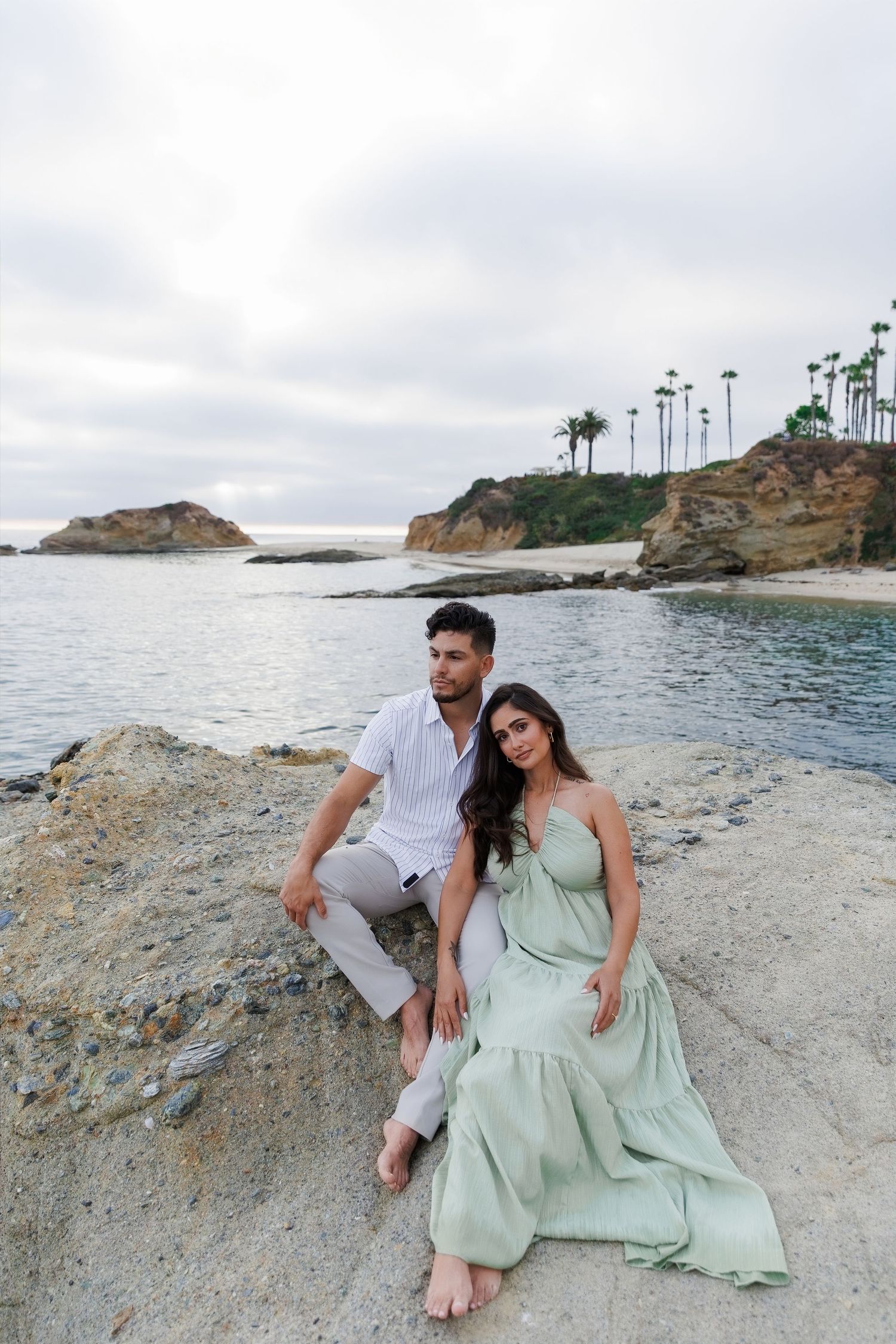 Couple sitting on coastal rocks in white and mint green outfits with palm trees and ocean cliffs in background.