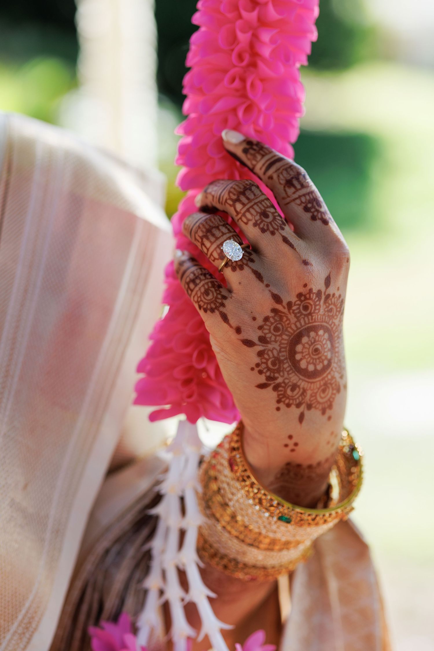 Bride's henna-adorned hand with gold bangles and diamond ring holding pink floral garland at Indian wedding.