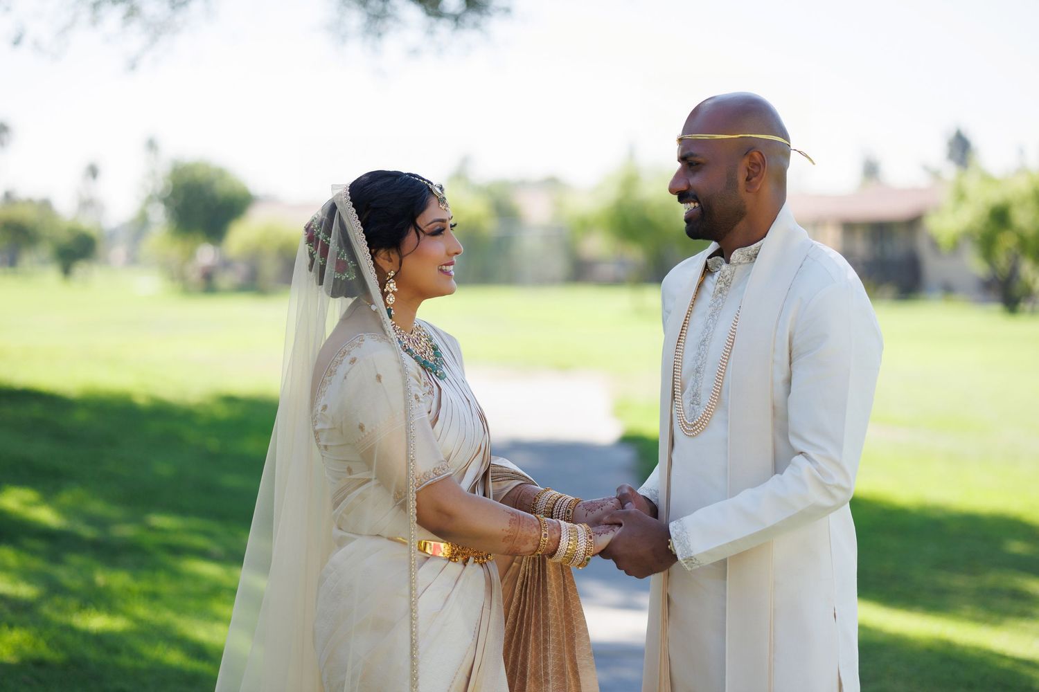 Indian bride and groom in traditional white wedding attire holding hands outdoors on lush green lawn.