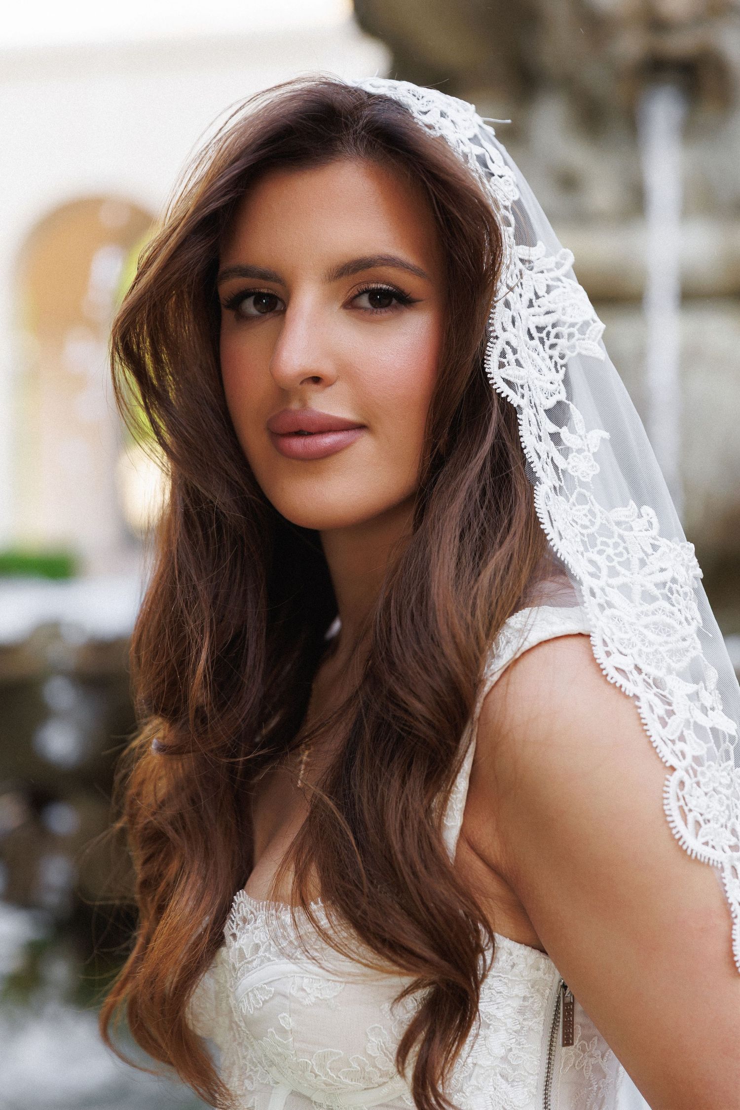 Elegant bride with long brunette hair wearing a lace veil and white dress, posing outdoors near a fountain.