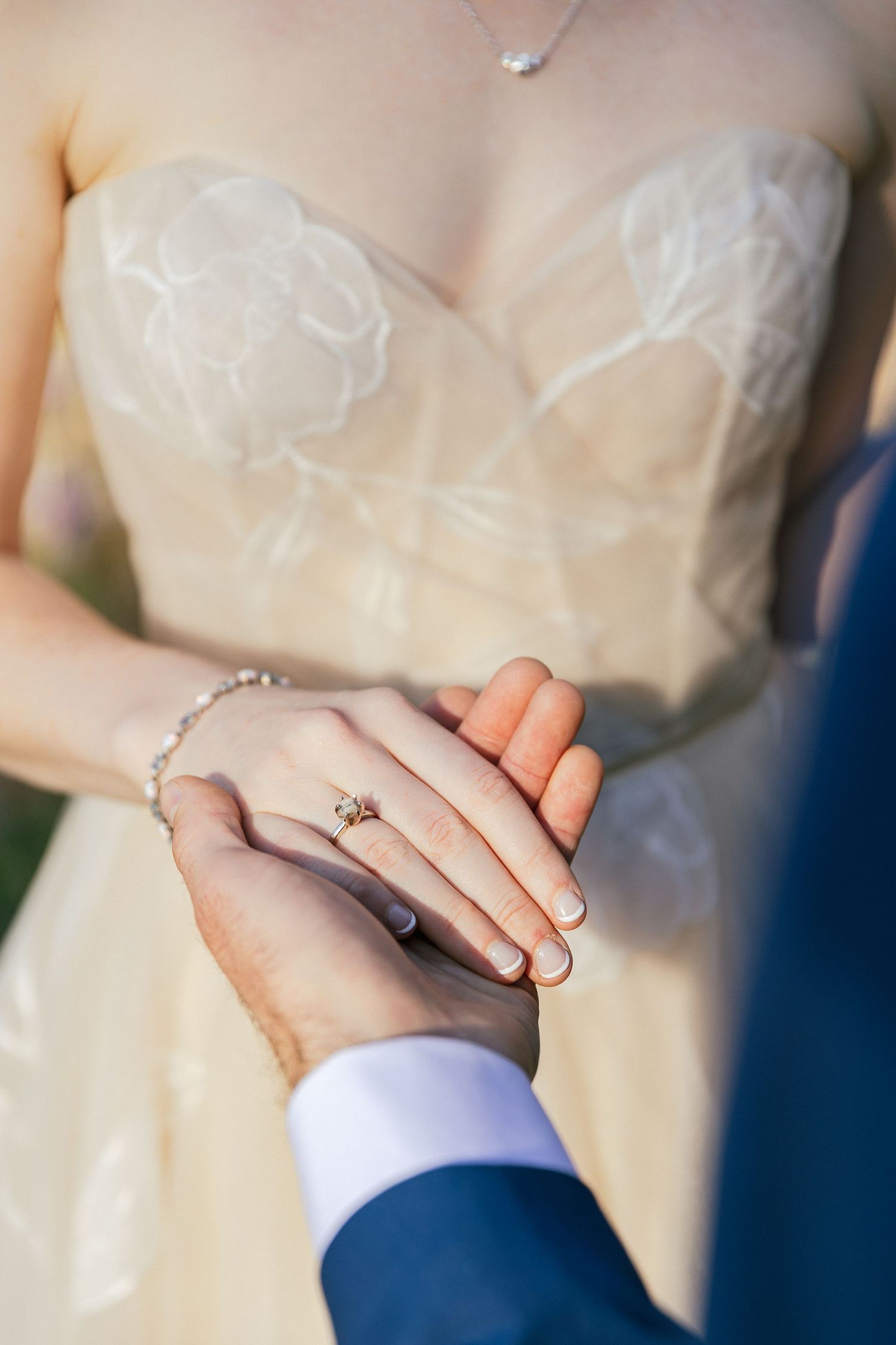 Close-up view of hands wearing wedding rings with white lace dress and navy blue suit in the background.