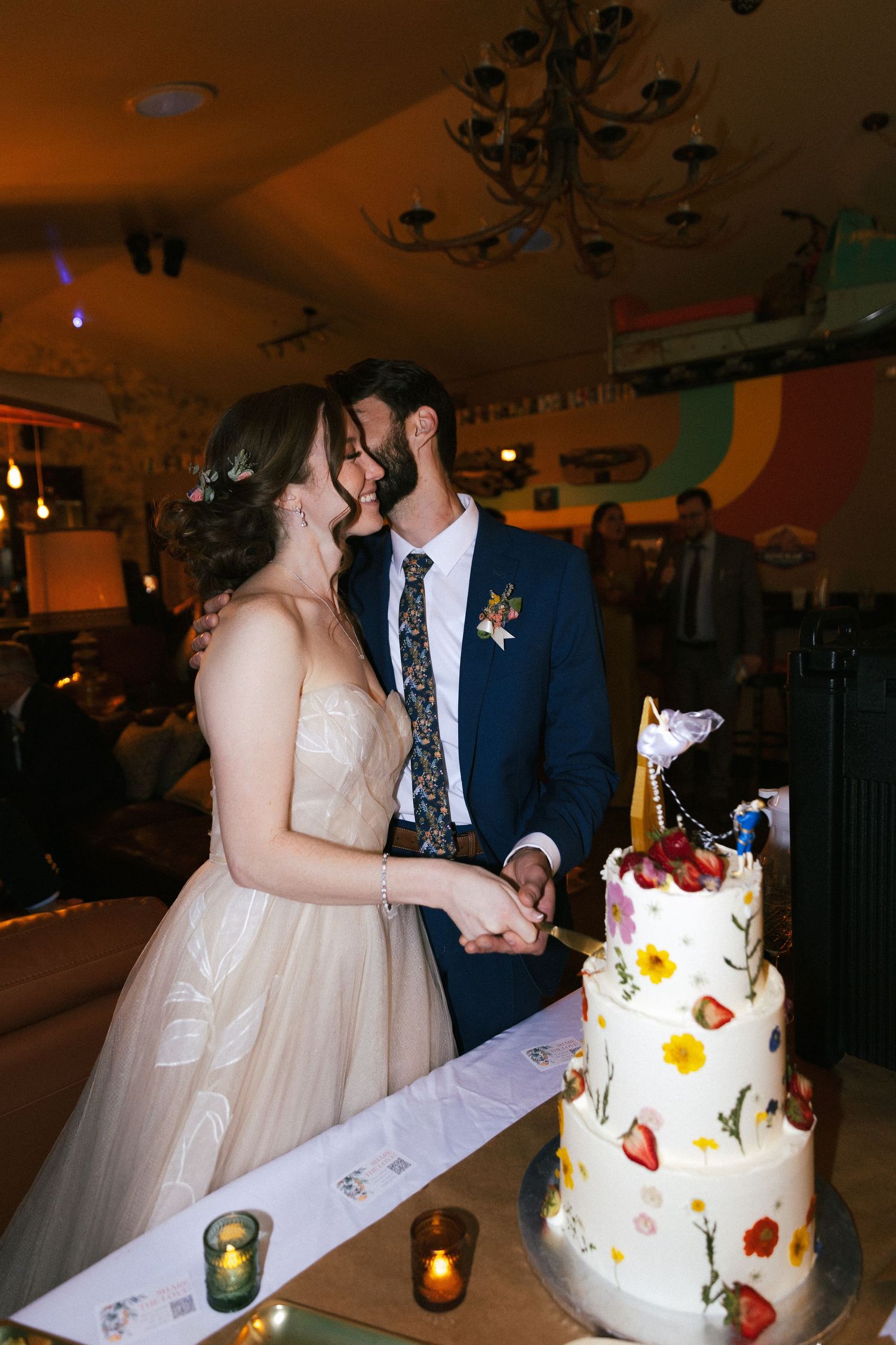 Bride and groom kissing while cutting a three-tier white wedding cake decorated with fresh flowers and strawberries.