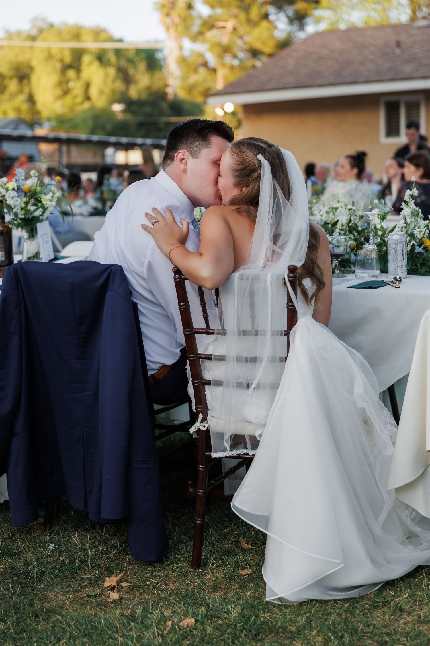 Bride and groom share a kiss at outdoor wedding reception table, bride wearing white gown and veil.