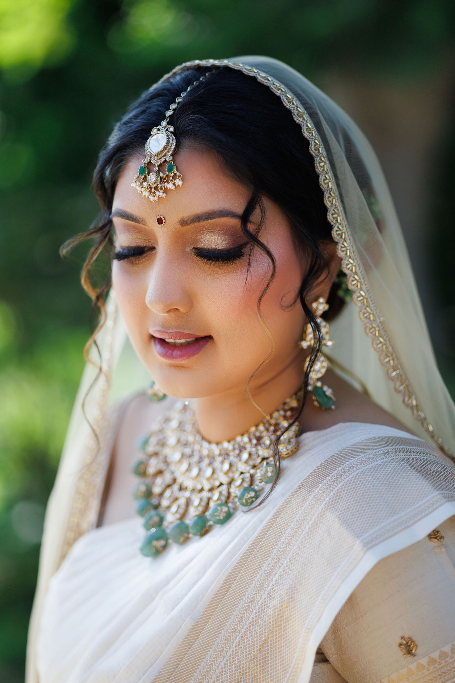 Indian bride in white saree with kundan jewelry, maang tikka, and veil, looking down gracefully outdoors.