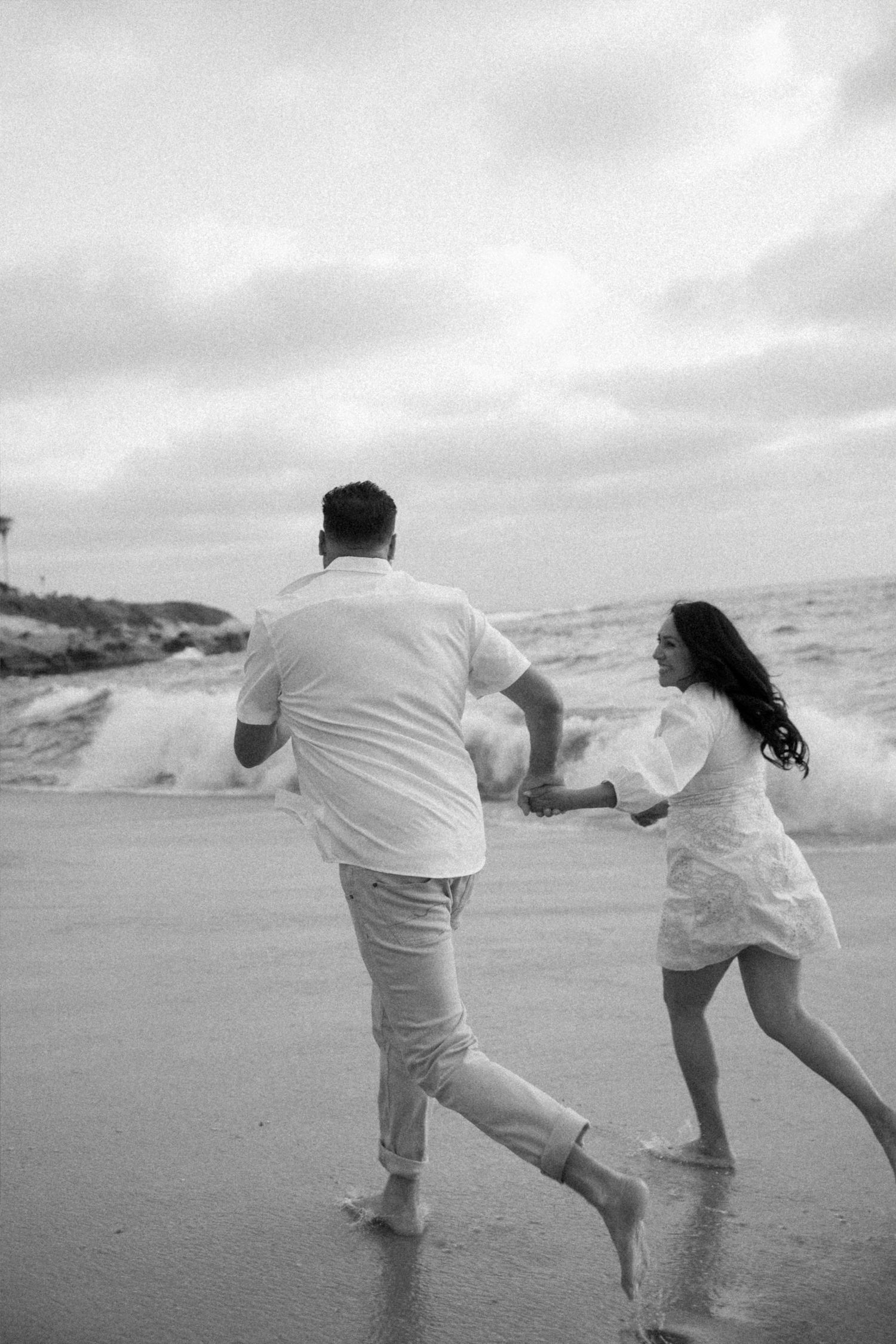 Black and white photo of a couple running barefoot on a beach, holding hands as waves crash behind them.