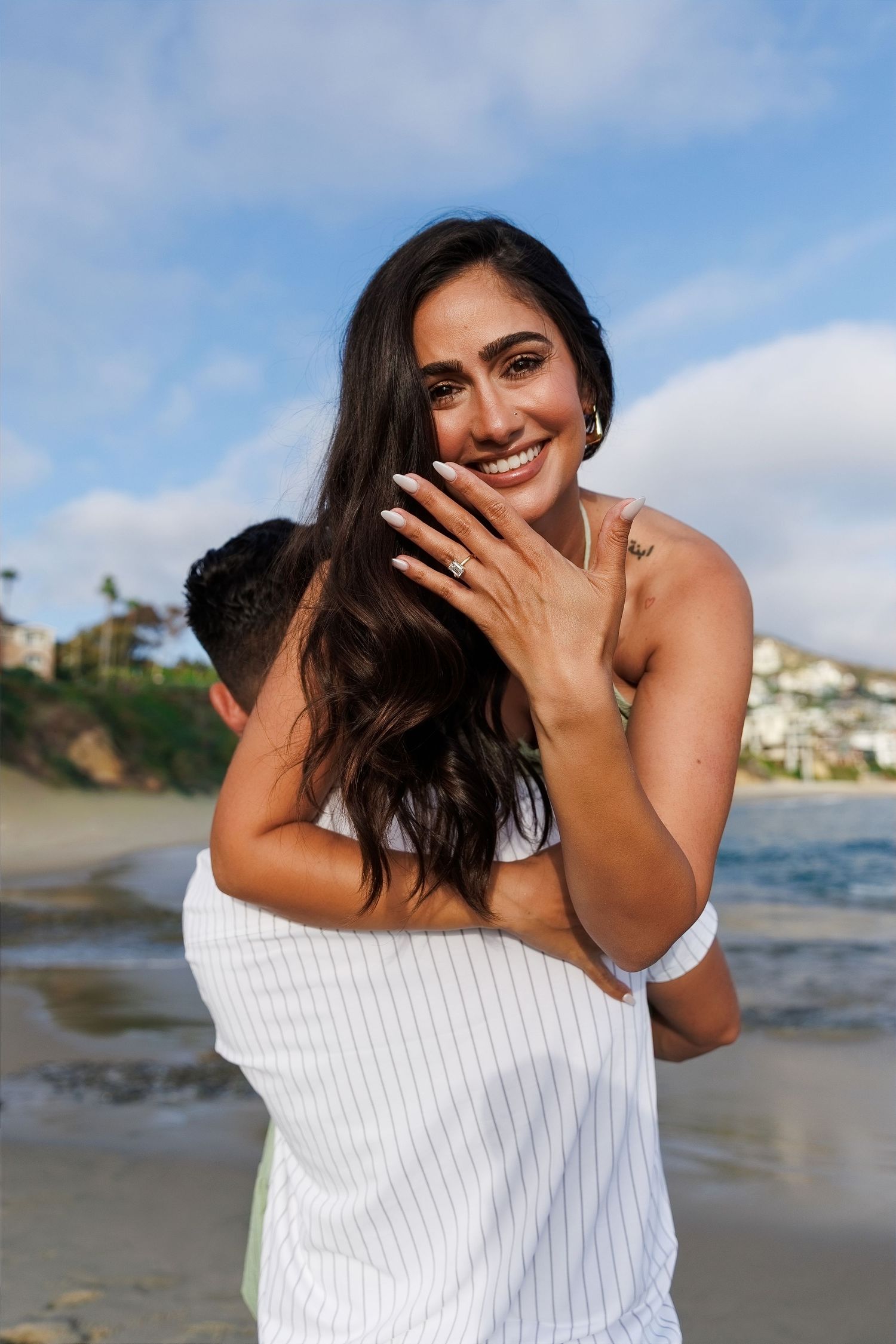 Smiling woman showing engagement ring on beach while being embraced by partner, with ocean and cliffs in background.