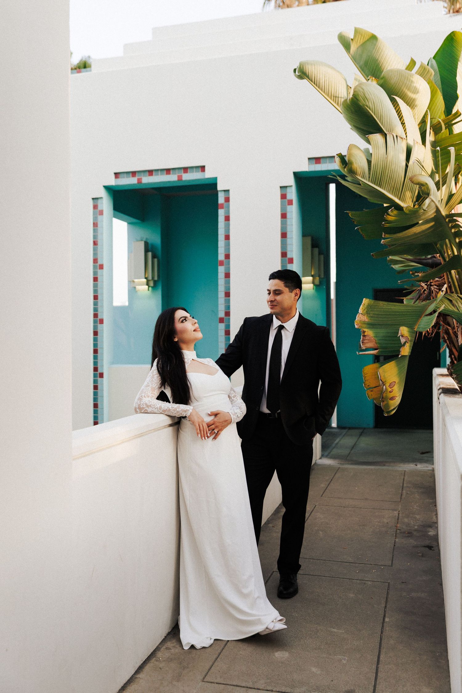 Bride in white gown and groom in black suit pose on a colorful teal and white venue balcony with tropical plants.