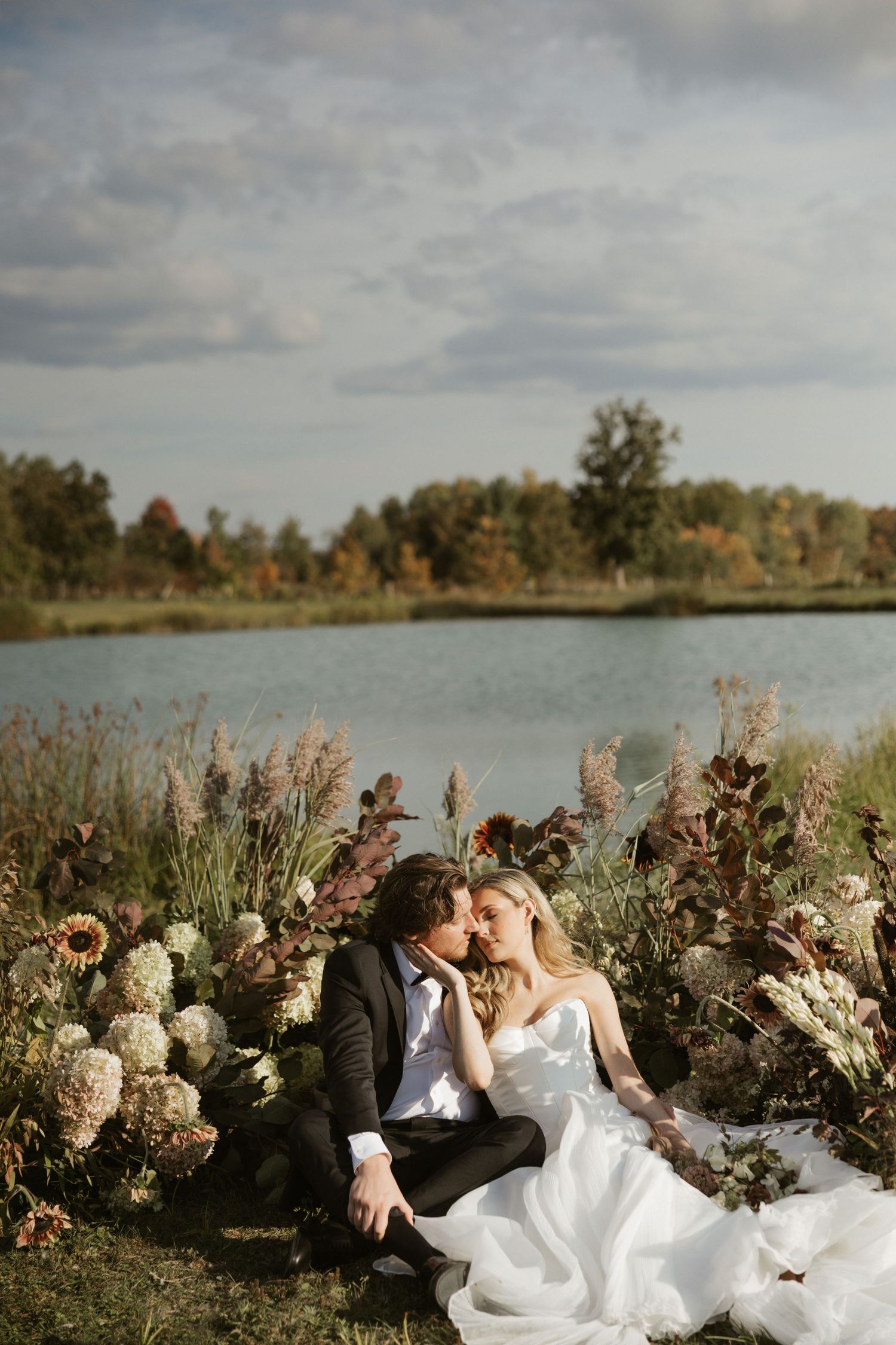 A couple relaxes in tall grass by a lake surrounded by autumn trees and wildflowers on a cloudy day.