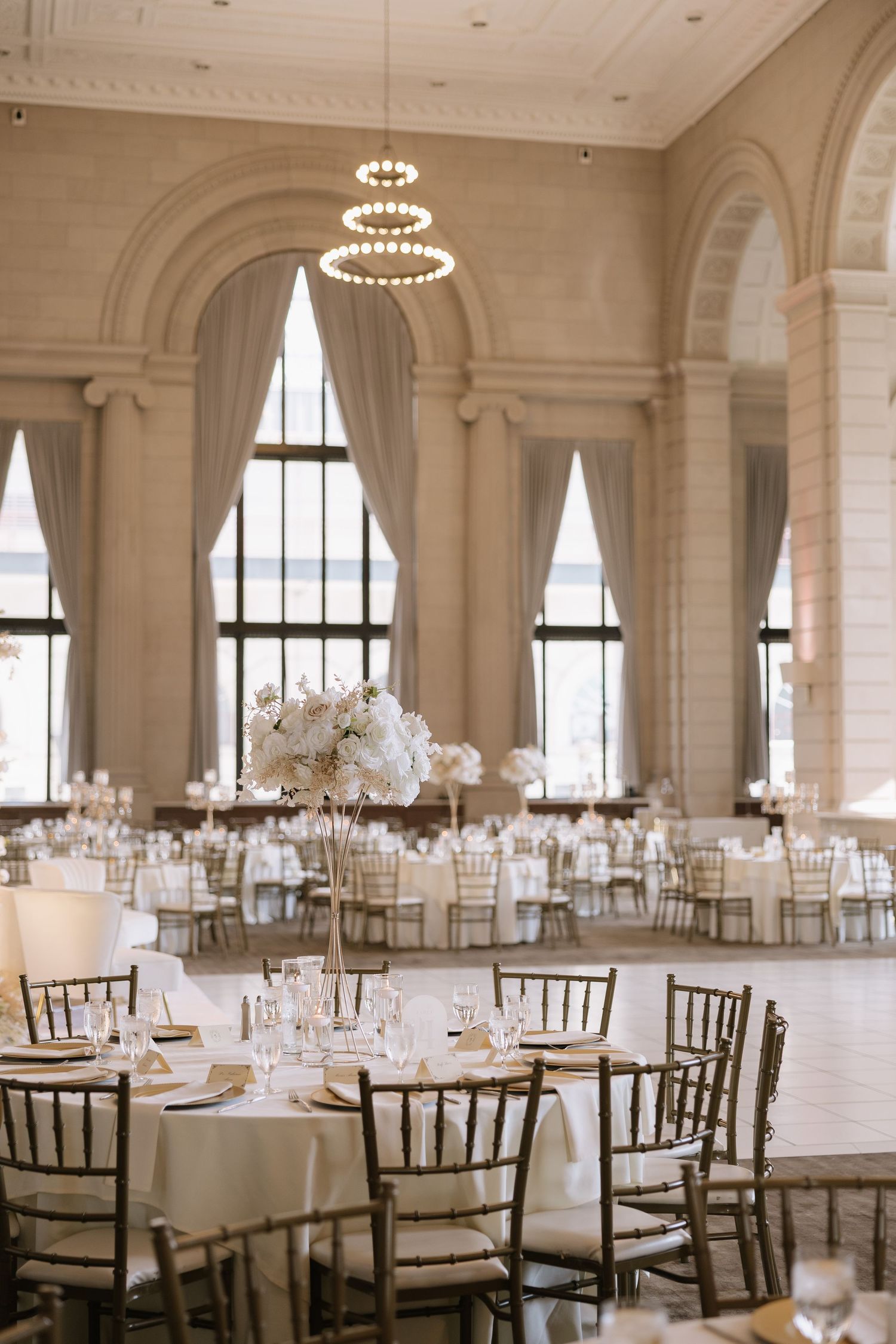 Elegant white ballroom with round tables, chiavari chairs, and crystal chandeliers set for a formal wedding reception.