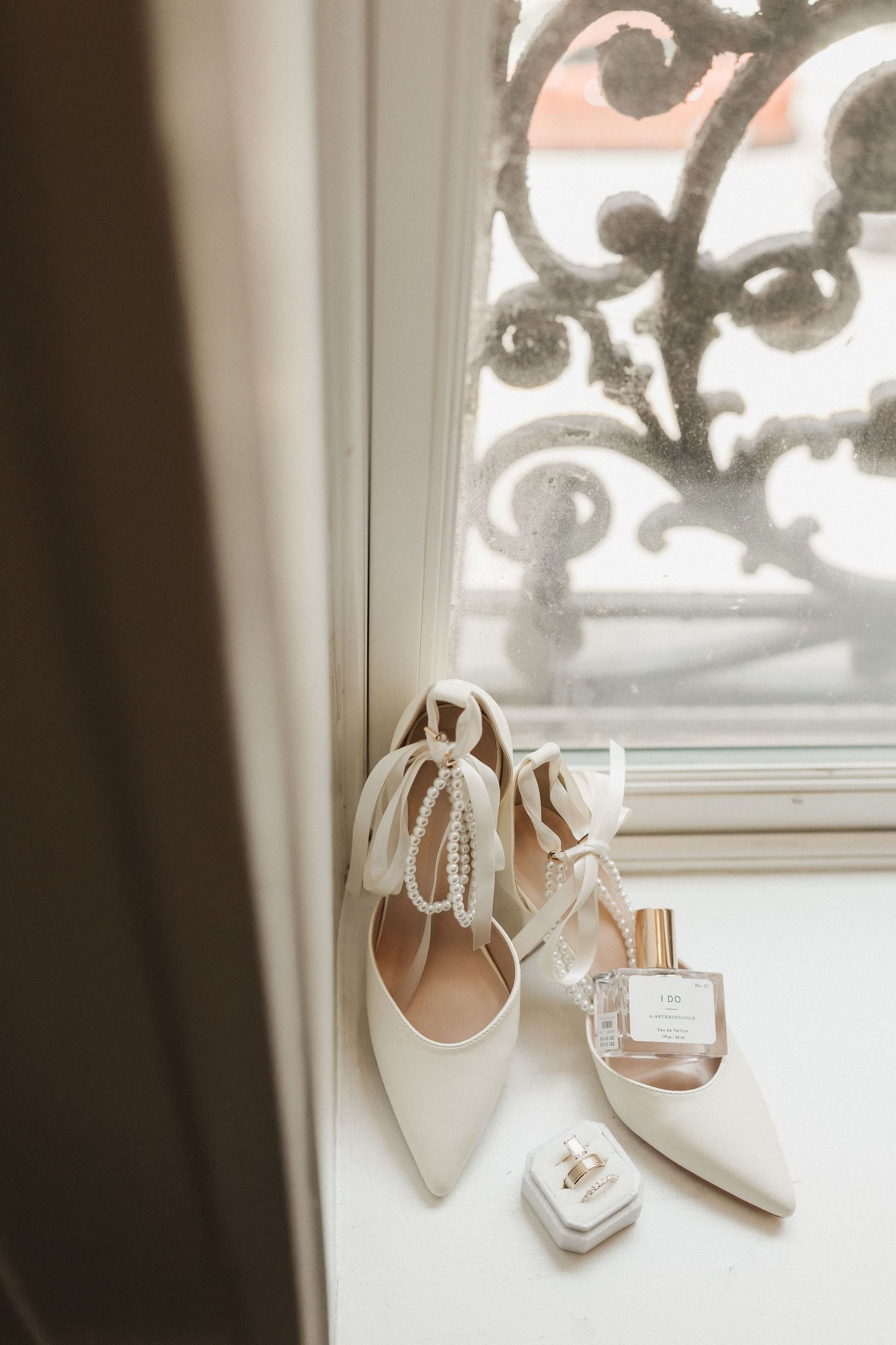 White bridal heels with pearl ankle ties beside perfume and ring box on windowsill with ornate ironwork backdrop.