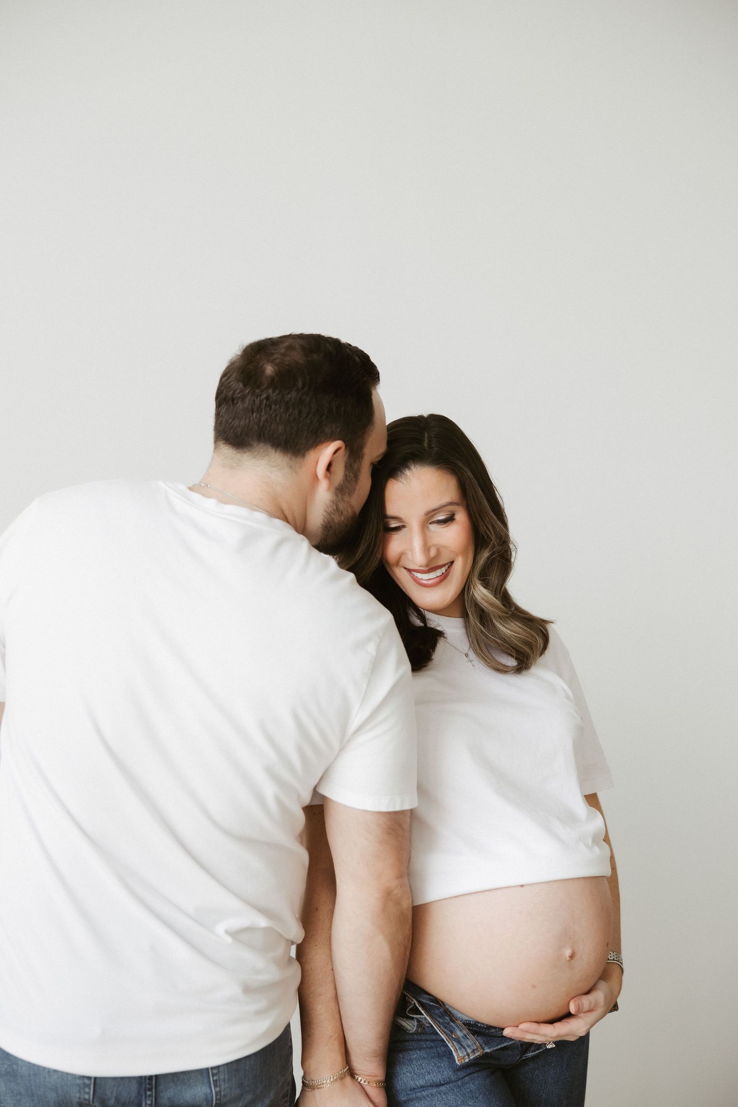 Smiling pregnant woman in white t-shirt holds her baby bump while partner whispers to her against white background.